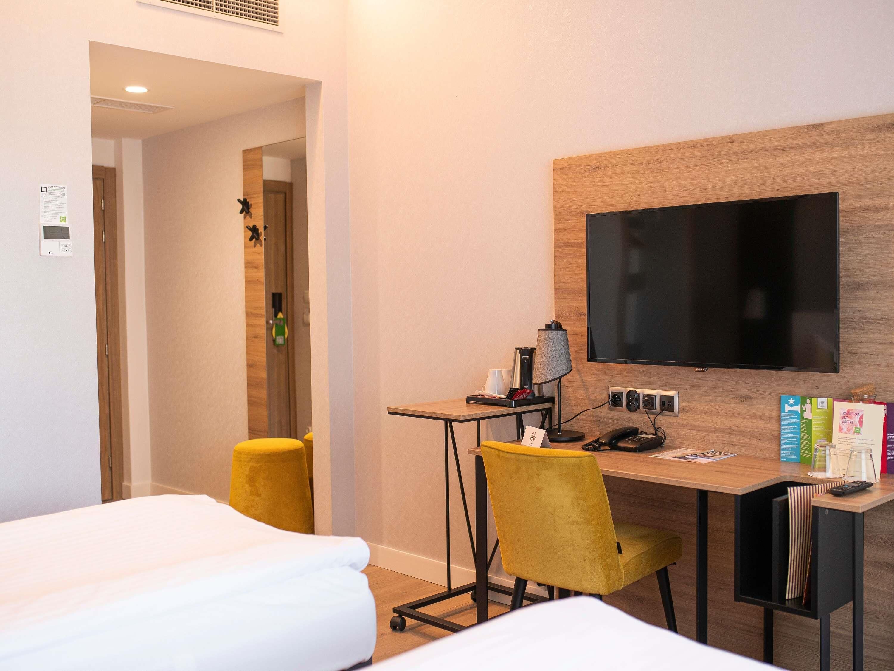 Ein Hotelzimmer mit einem Bett, einem Schreibtisch mit Stuhl, einem Fernseher und einem kleinen Tisch mit Lampe.