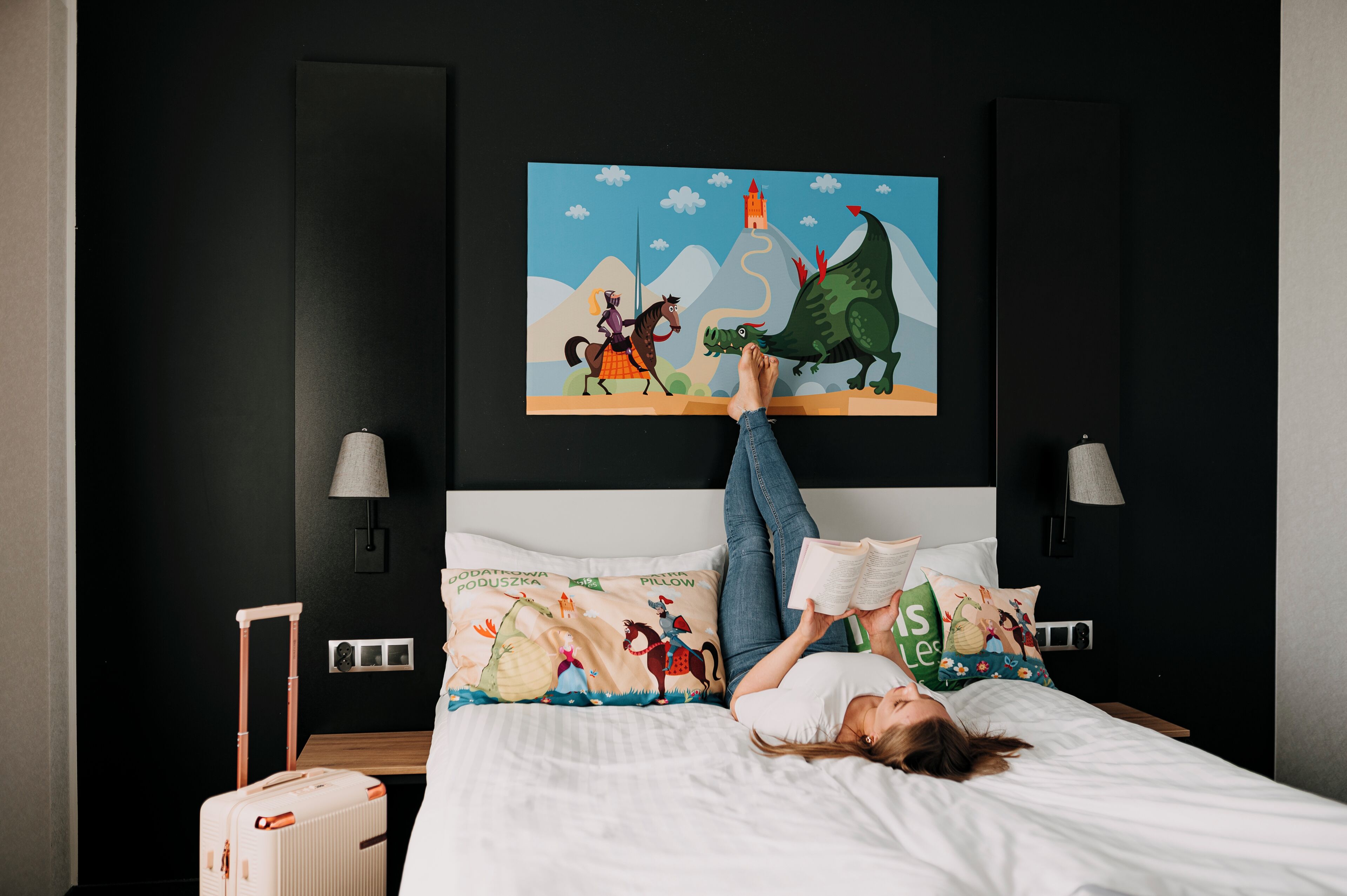Una cama con un cojín colorido, una maleta y un cuadro en la pared que muestra un caballero, un caballo y un dragón.