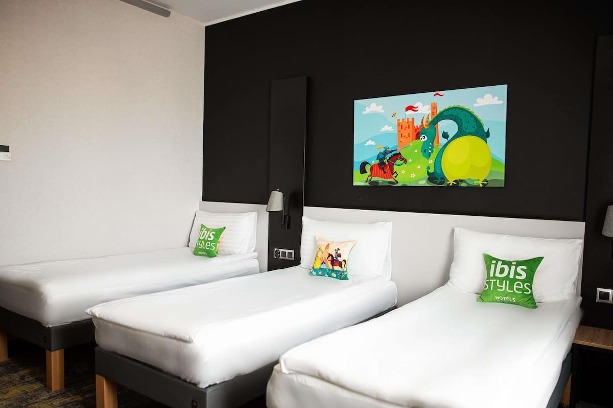 A hotel room with three single beds, a black headboard, a wall-mounted lamp, and a colorful painting above the beds.