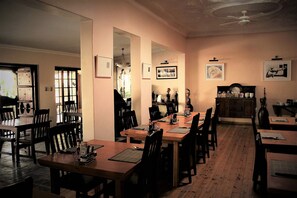 A dining area with wooden tables and chairs, a ceiling fan, and framed pictures on the walls. - African Roots Guest House (Polokwane)