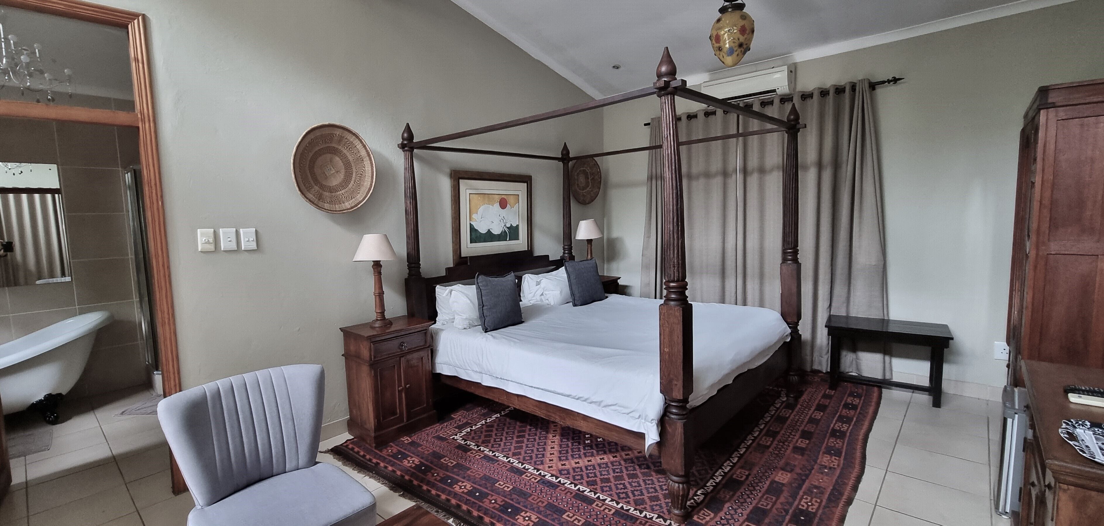 A four-poster bed with a wooden frame, a bedside table with a lamp, a chair, a patterned rug, and a large mirror.