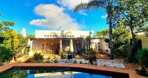 A pool with a clear view of the surrounding garden and a building with a patio area. - African Roots Guest House (Polokwane)