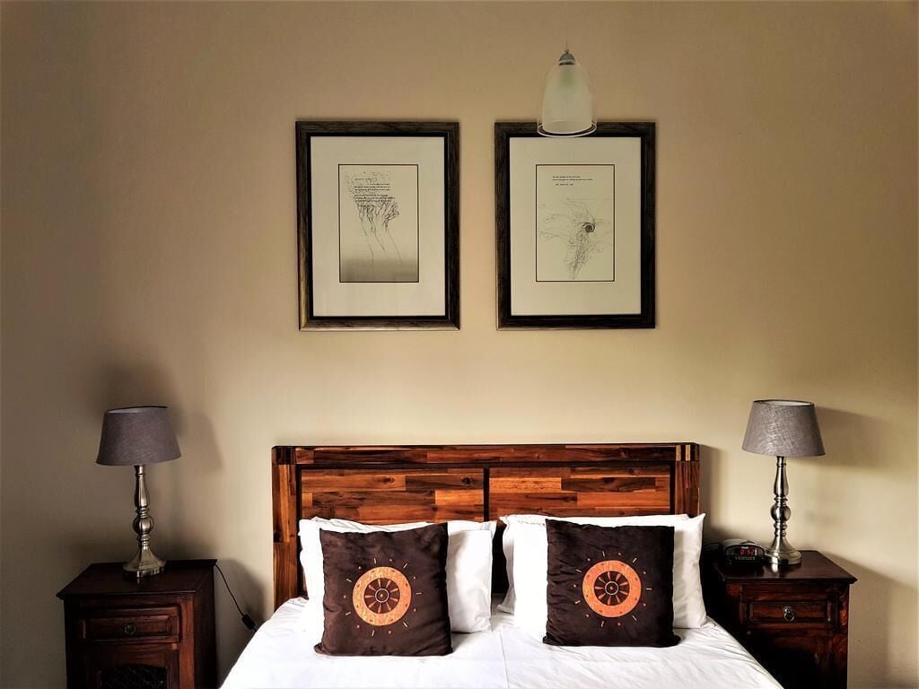 A hotel room with a wooden headboard, two bedside tables with lamps, a bed with white bedding and two decorative pillows, and two framed artworks on the wall.