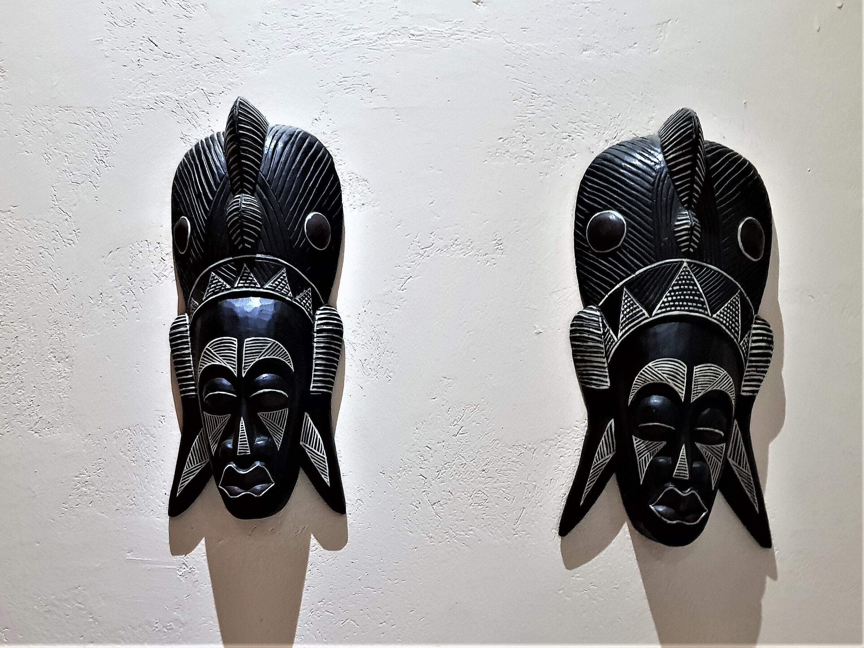Two black wooden masks with geometric patterns and circular eyes on a plain wall.