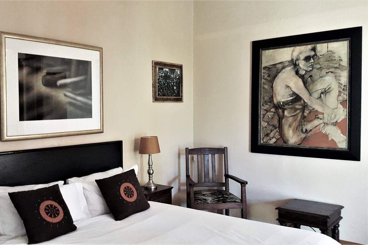 A hotel room with a large bed, two bedside tables with lamps, a chair, and three framed artworks on the wall.
