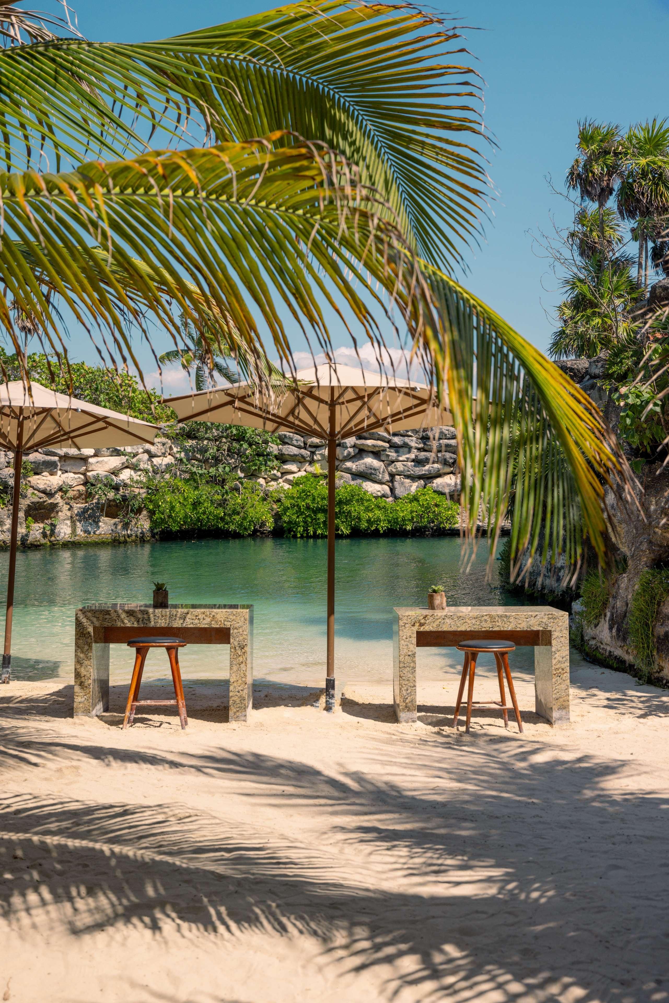 A beachside seating area with two stone tables and chairs, a palm tree, and a view of a calm body of water and rocky landscape.
