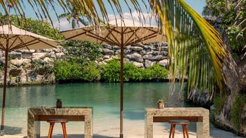 A beachside seating area with two stone tables and chairs, a palm tree, and a view of a calm body of water and rocky landscape.