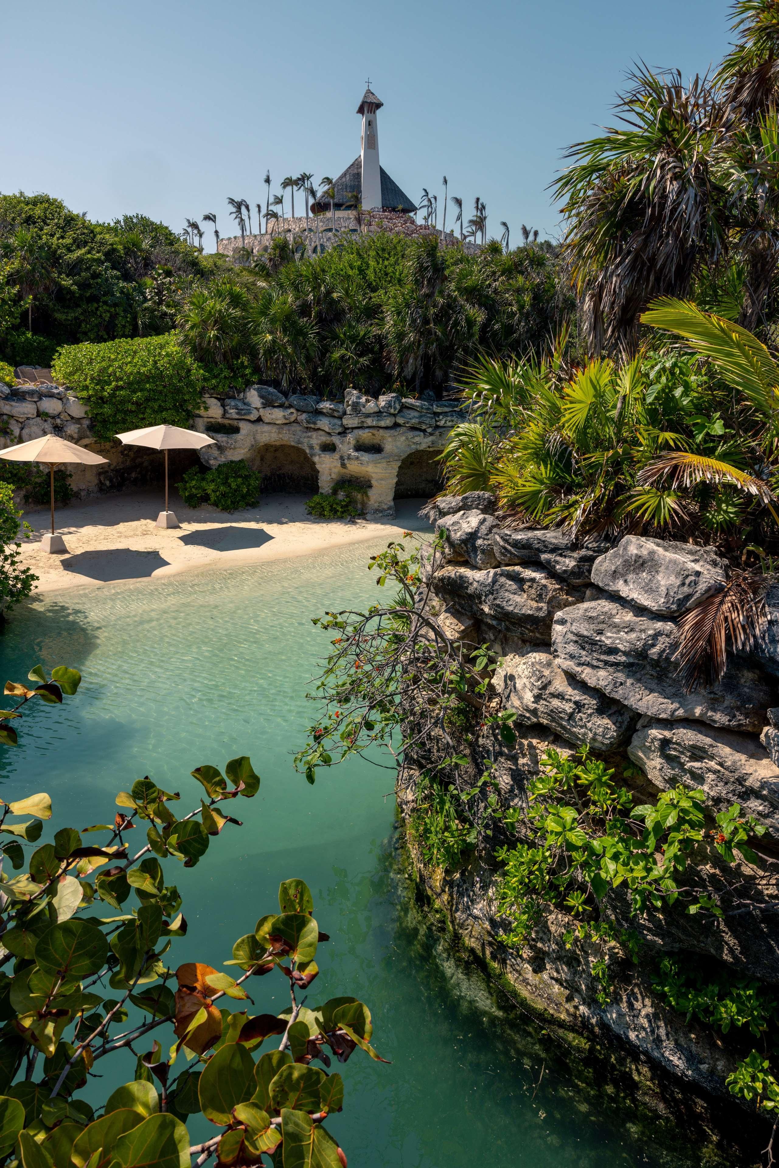 A clear pool surrounded by rocks and greenery, with a stone wall and palm trees in the background.