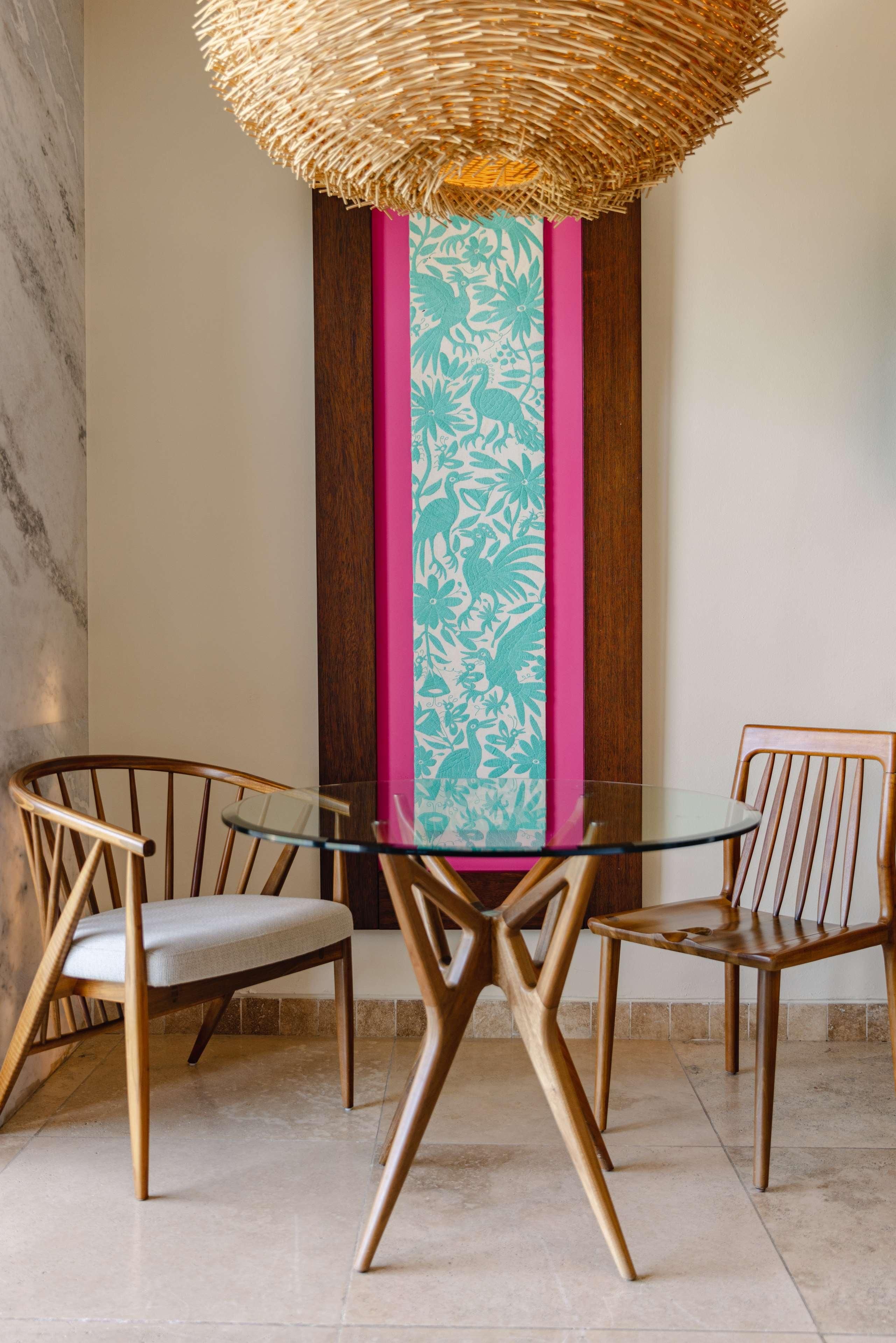 A dining area with a glass table and two wooden chairs, a woven ceiling light fixture, and a wall with a vertical wooden panel and a pink and teal patterned panel.
