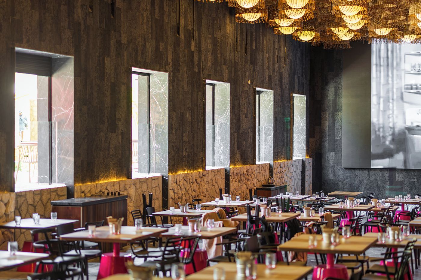 A restaurant with multiple tables and chairs, large windows, and a stone wall.