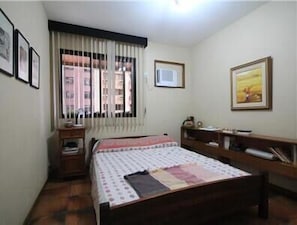 A small, neatly arranged bedroom with a single bed, a bedside table, a chair, and a window with curtains.