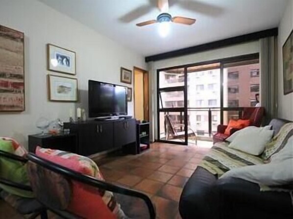 A living room with a television on a cabinet, a ceiling fan, and a large window with a view of buildings.