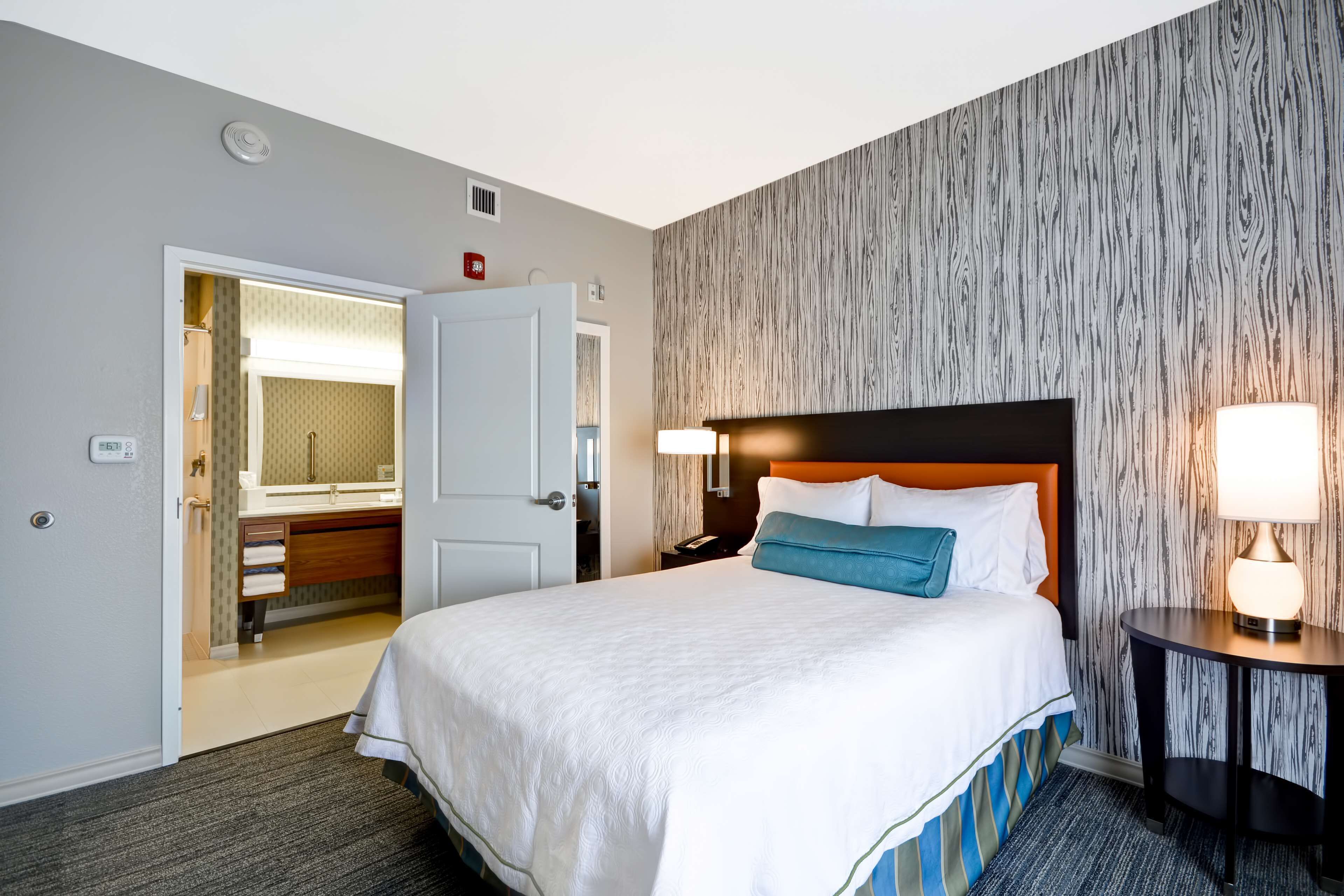 Habitación de hotel con cama, mesita de noche, lámpara, espejo y baño visible a través de una puerta abierta.