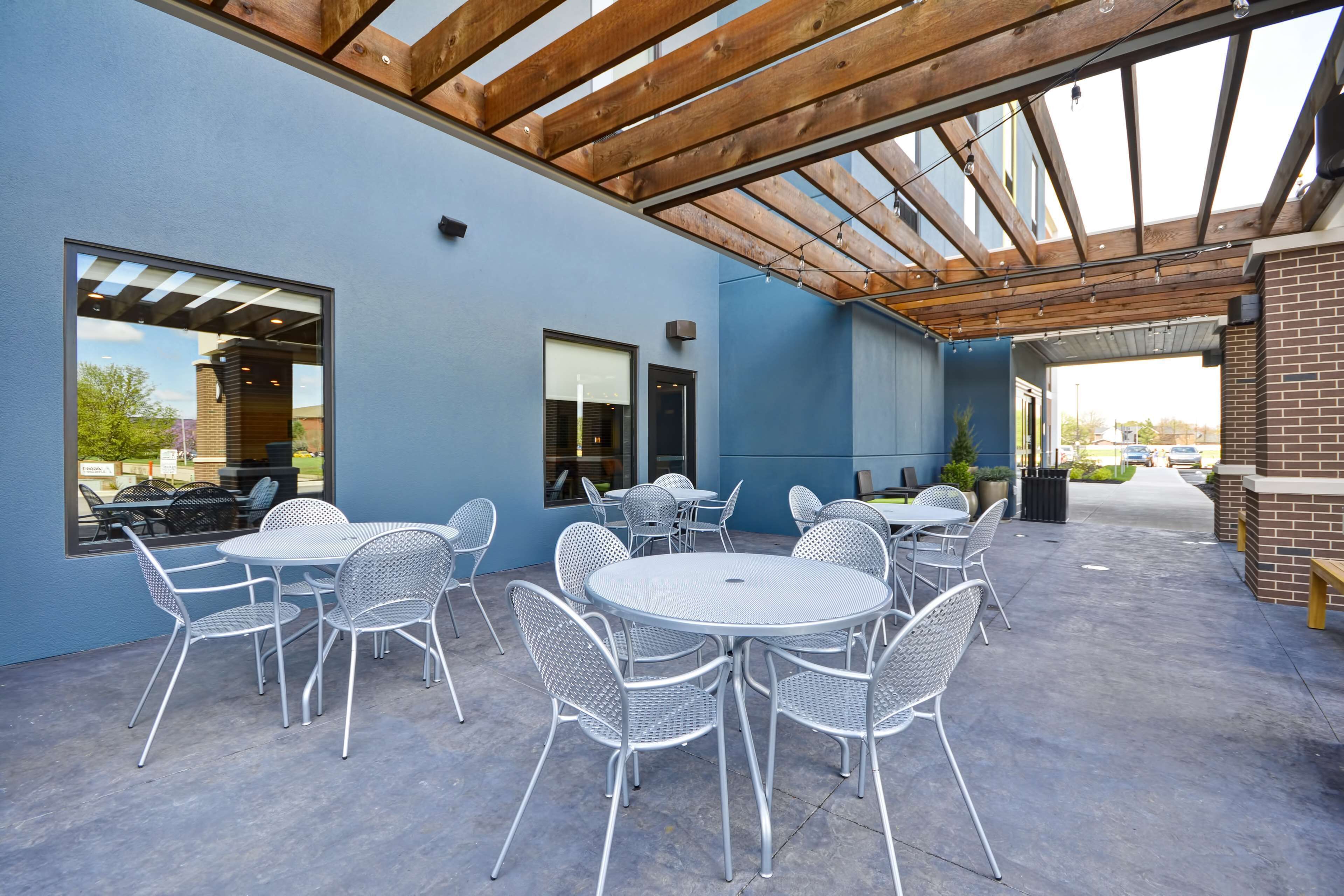 Un patio al aire libre con mesas redondas y sillas de metal, una estructura de techo de madera y ventanas grandes.