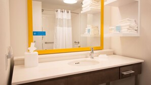 A hotel bathroom with a white sink, a mirror with a yellow frame, a towel rack, and a shelf with toiletries.