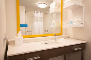 A hotel bathroom with a white sink, a mirror with a yellow frame, a towel rack, and a shelf with toiletries.