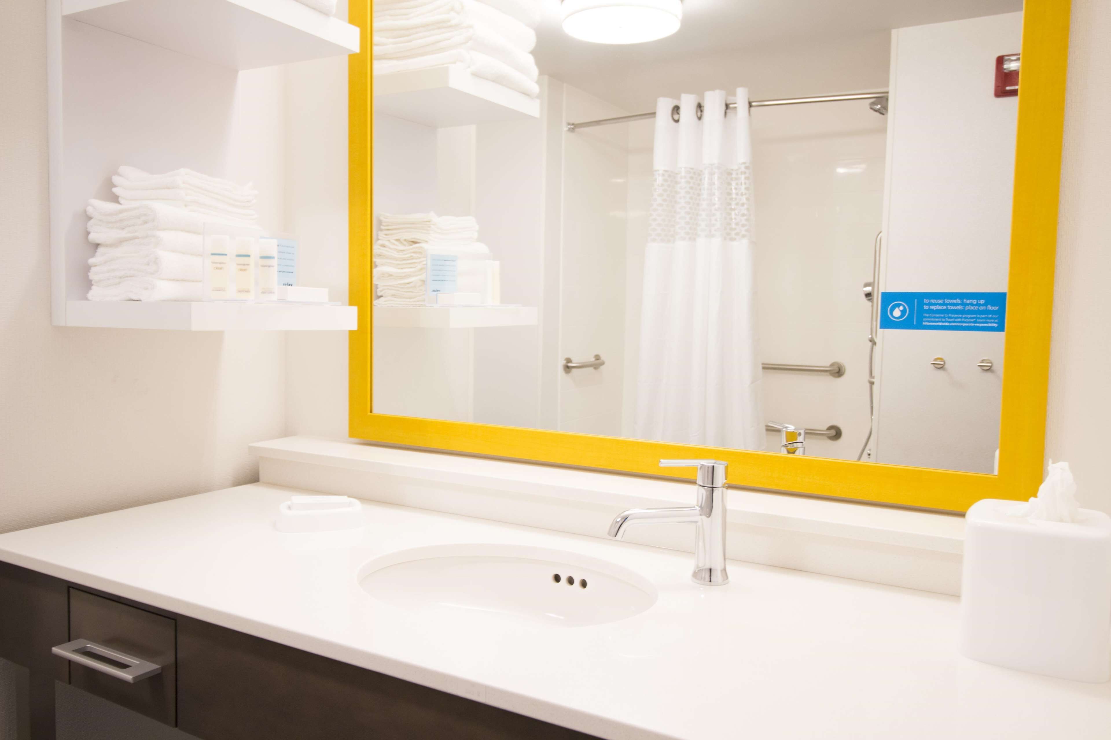A modern bathroom with a white sink, a large mirror with a yellow frame, and neatly folded towels on a shelf.