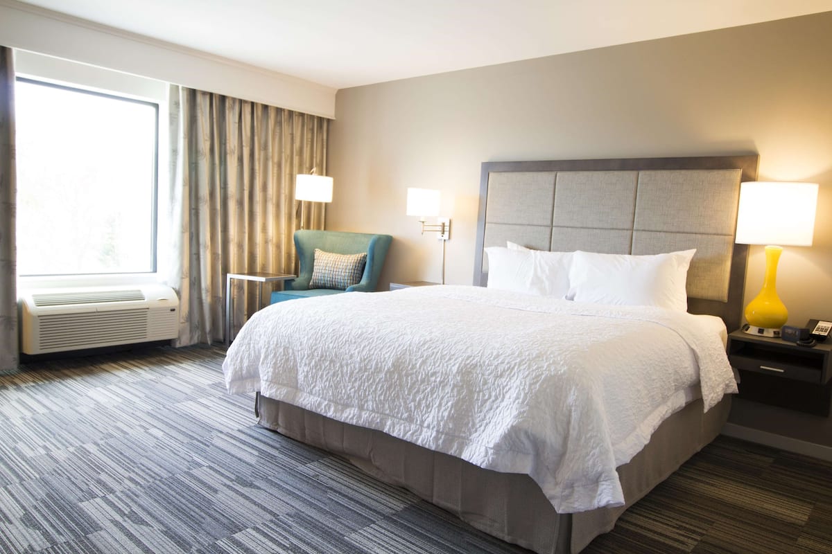 Hotelkamer met een groot bed, een stoel, een nachtkastje met lamp, een raam met gordijnen, en een airconditioning.