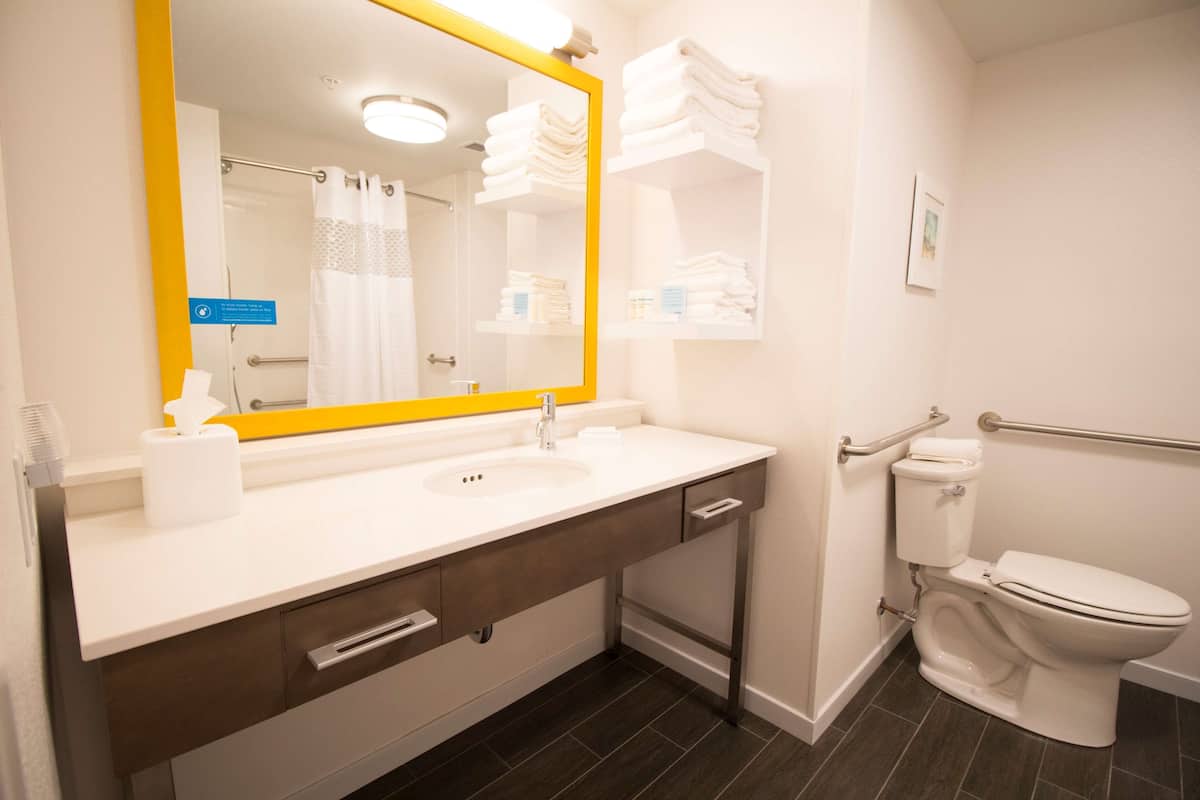 Een badkamer met een wit wastafel, spiegel en toilet. Handdoeken liggen op een plank gestapeld en er is een handgreep naast het toilet.
