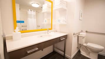 A bathroom with a white sink, mirror, and toilet. There are towels stacked on a shelf and a handrail next to the toilet.