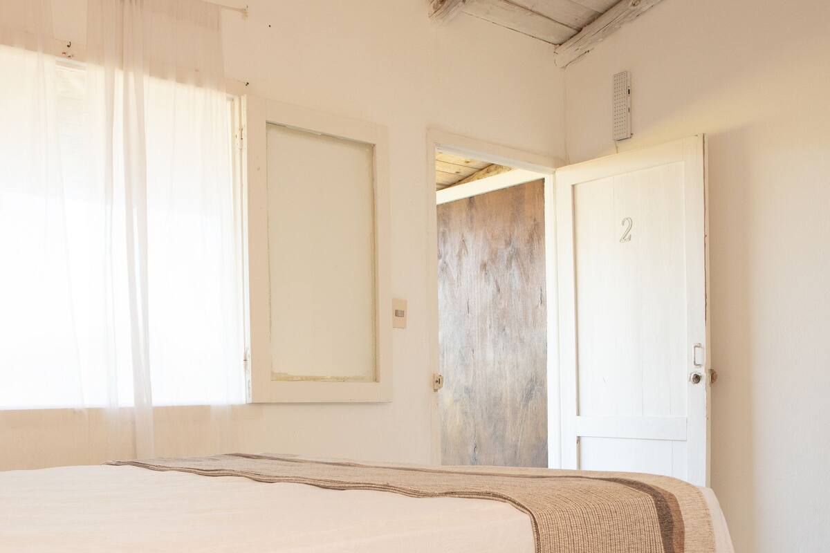 A bedroom with a large bed, a door with the number 2, and a window with sheer curtains.