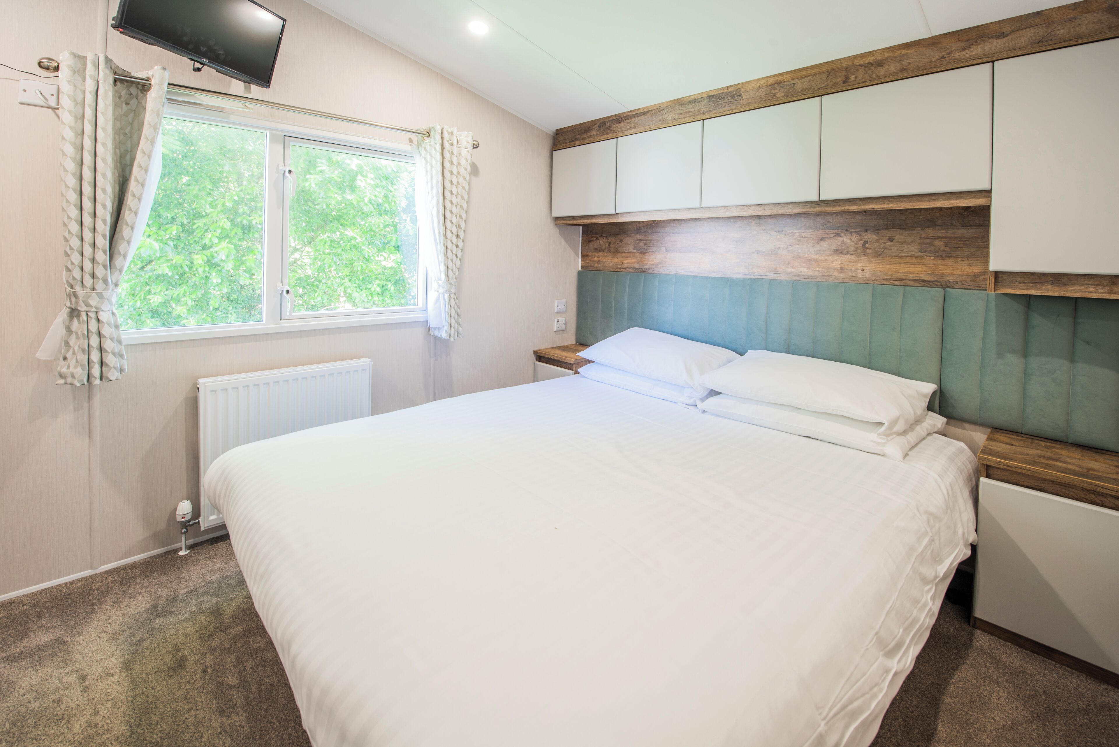 A bedroom with a bed, a window, a radiator, and a television mounted on the wall.