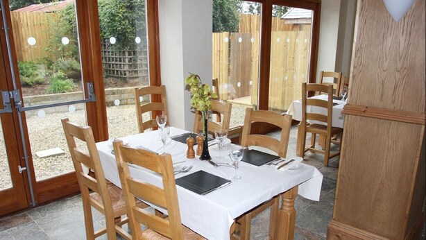 A dining area with a table set for four, surrounded by wooden chairs, with a view of a garden through large glass doors. - Cottage Lea's Country Hotel & Restaurant (Pickering)
