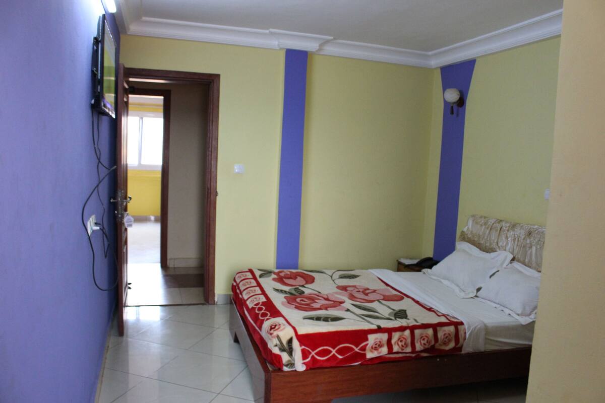 A bedroom with a bed, a door leading to another room, and a wall-mounted light fixture.
