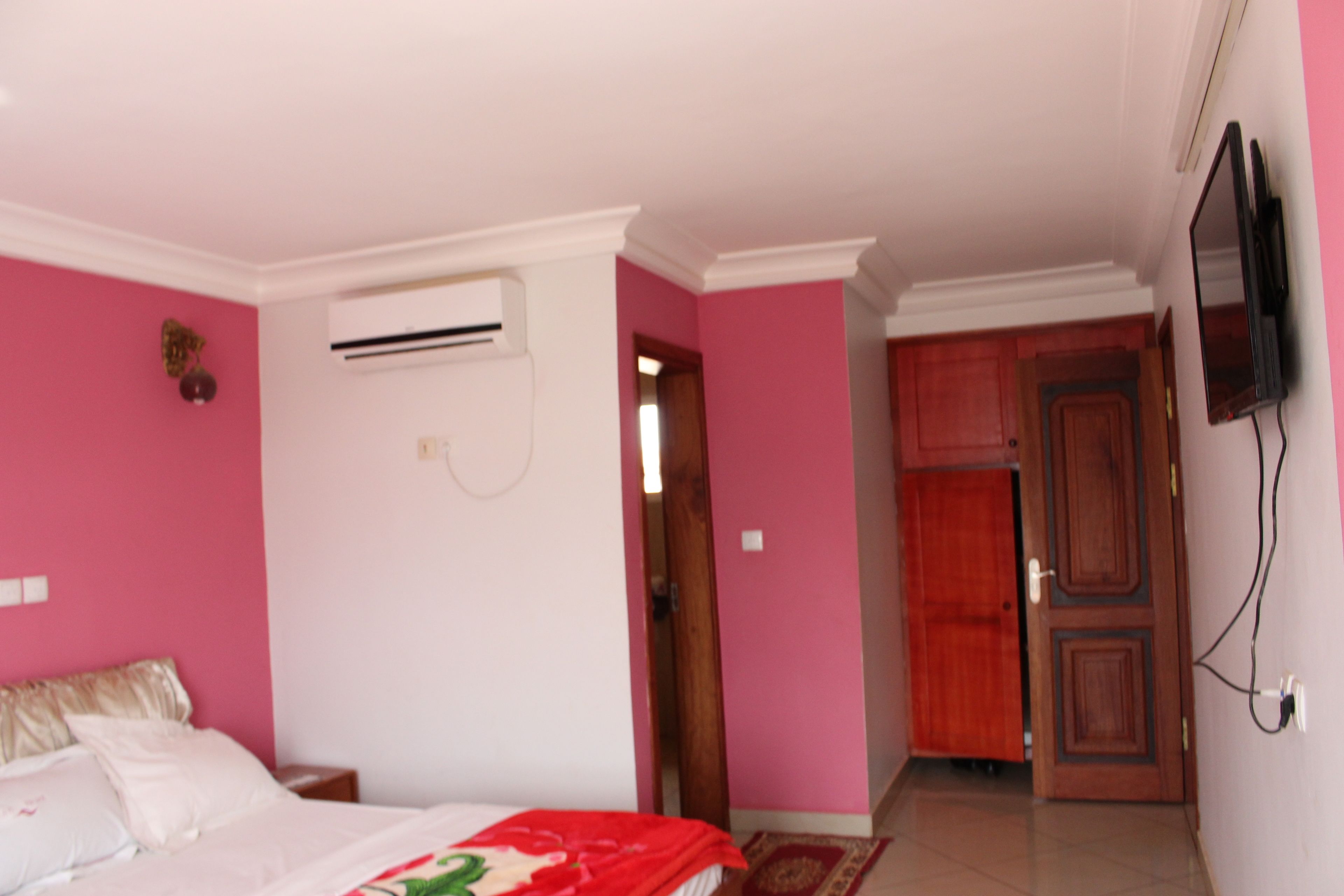 A bedroom with pink walls, a bed with white bedding, an air conditioning unit, a wooden door, and a television mounted on the wall.