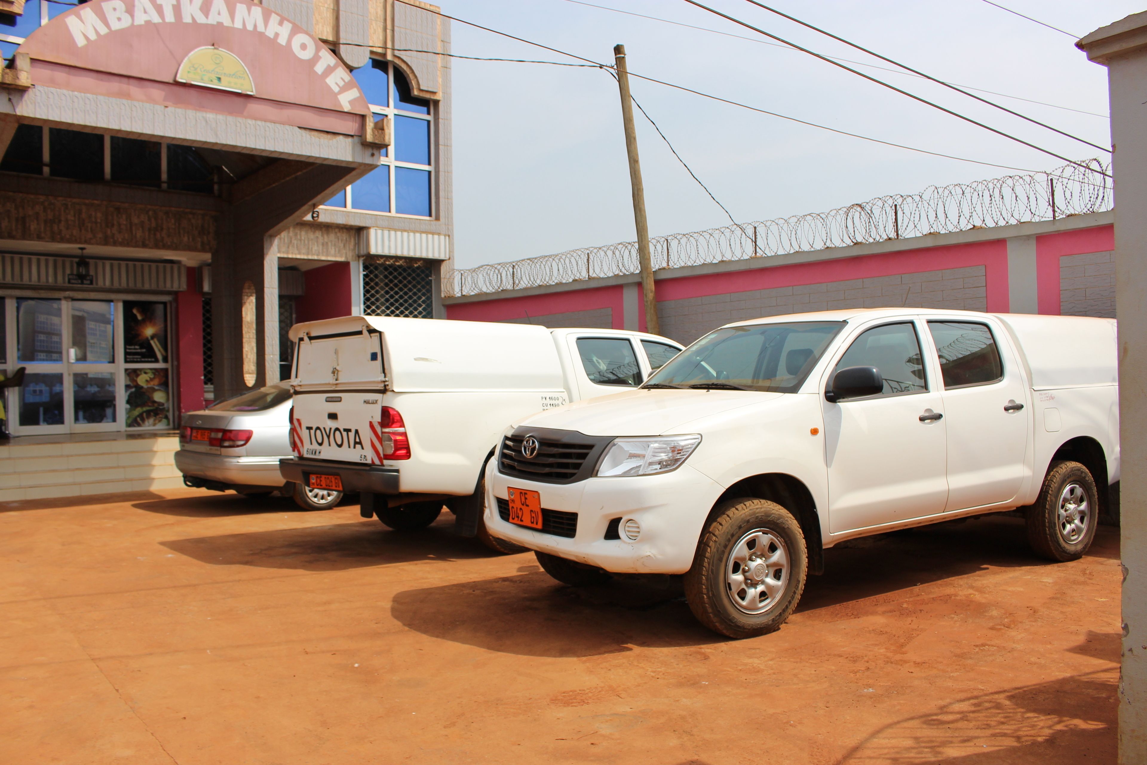 A hotel with a sign reading "MBATKAMHOTEL" and two white Toyota pickup trucks parked in front.