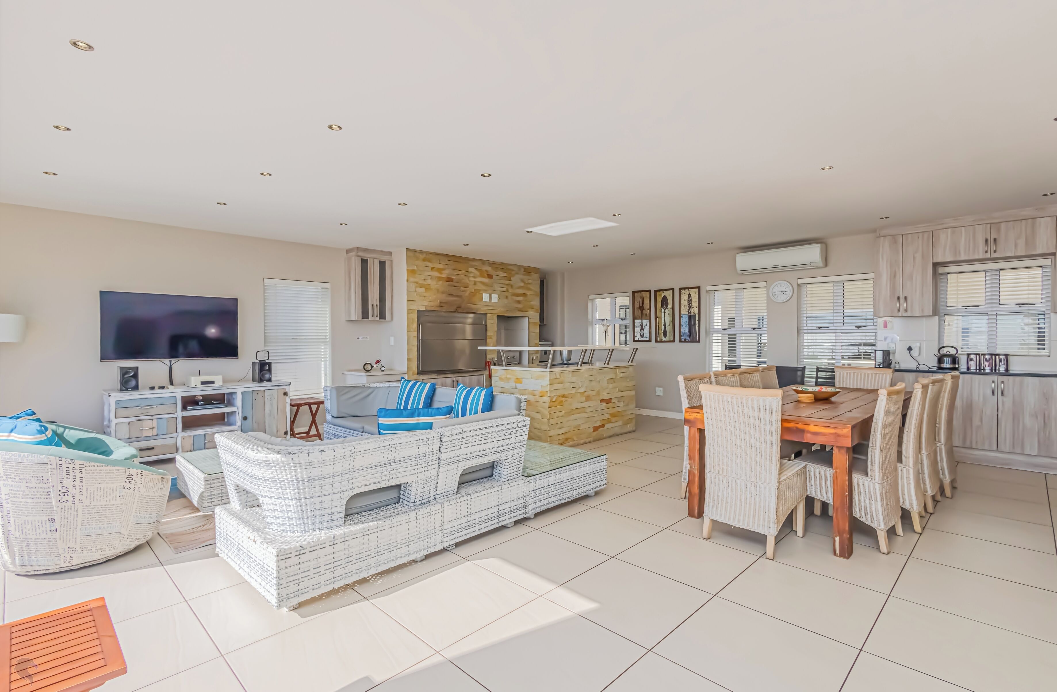 Een ruime woonkamer met een grote televisie, rieten meubels, een keuken met houten kastjes en een eettafel met stoelen.