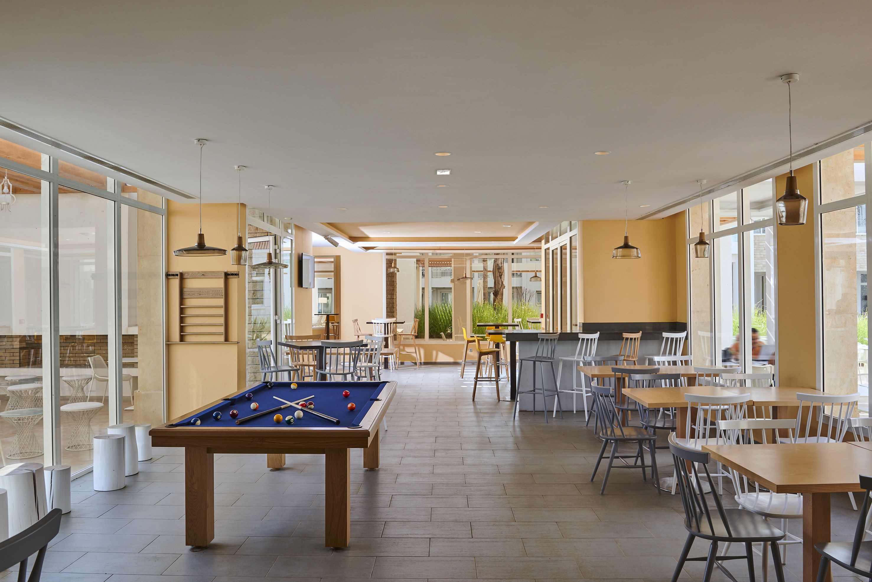 A pool table in a bright, open space with tables and chairs, large windows, and a bar area.