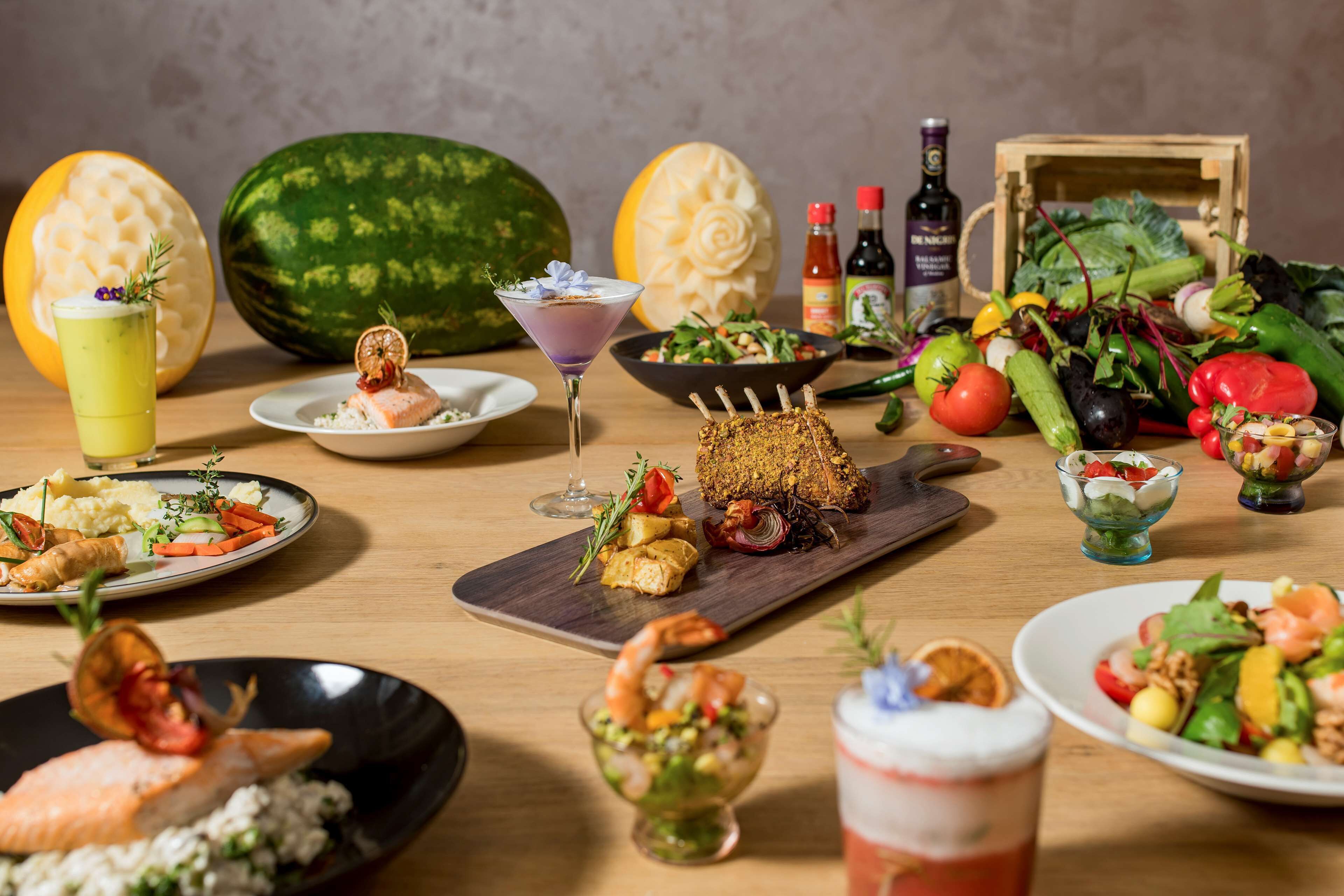 A variety of dishes and drinks on a wooden table, including a watermelon, salad, and seafood.