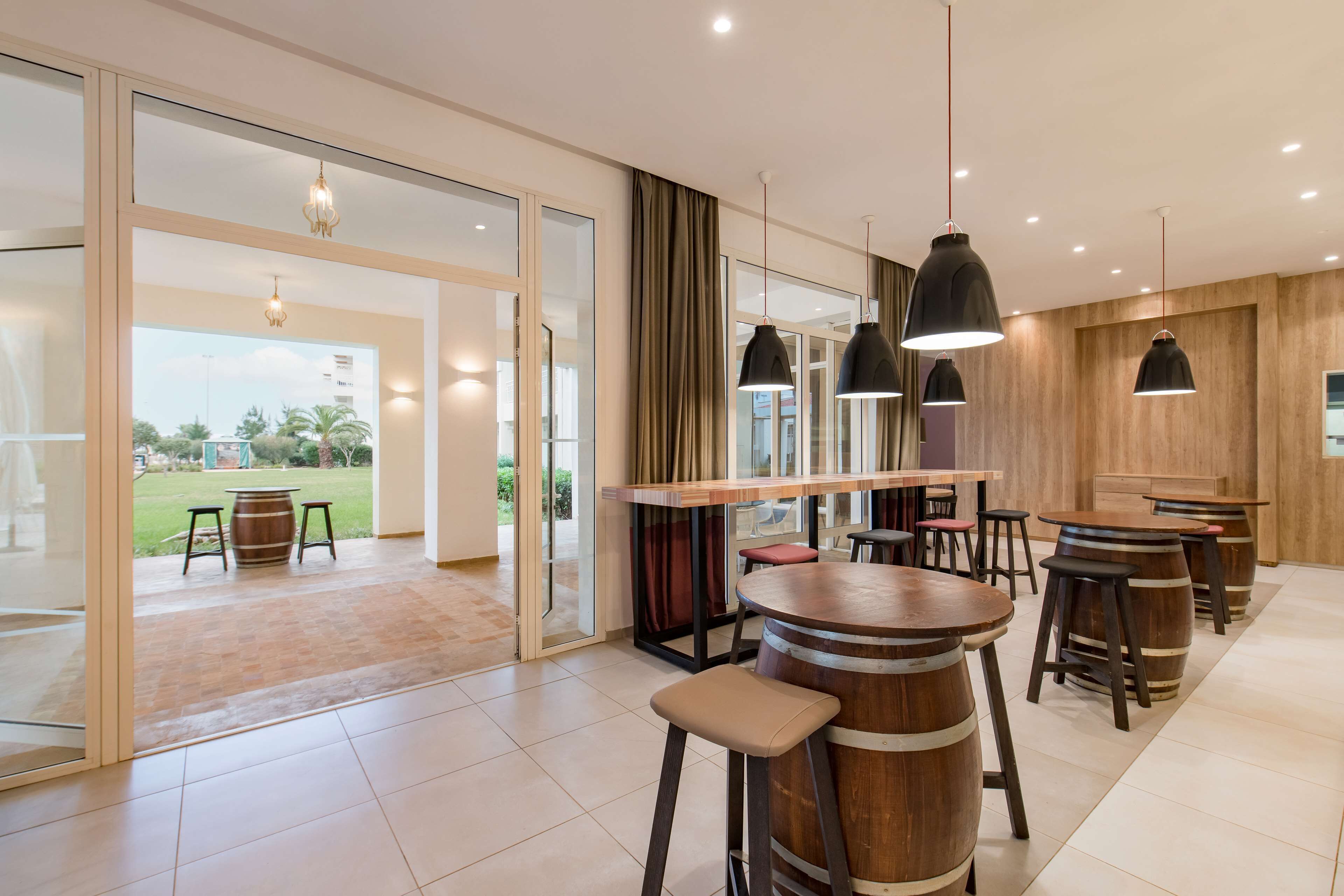 Zona de bar moderna con barriles de madera como taburetes, grandes lámparas colgantes y un puerta de cristal que da a un patio al aire libre.