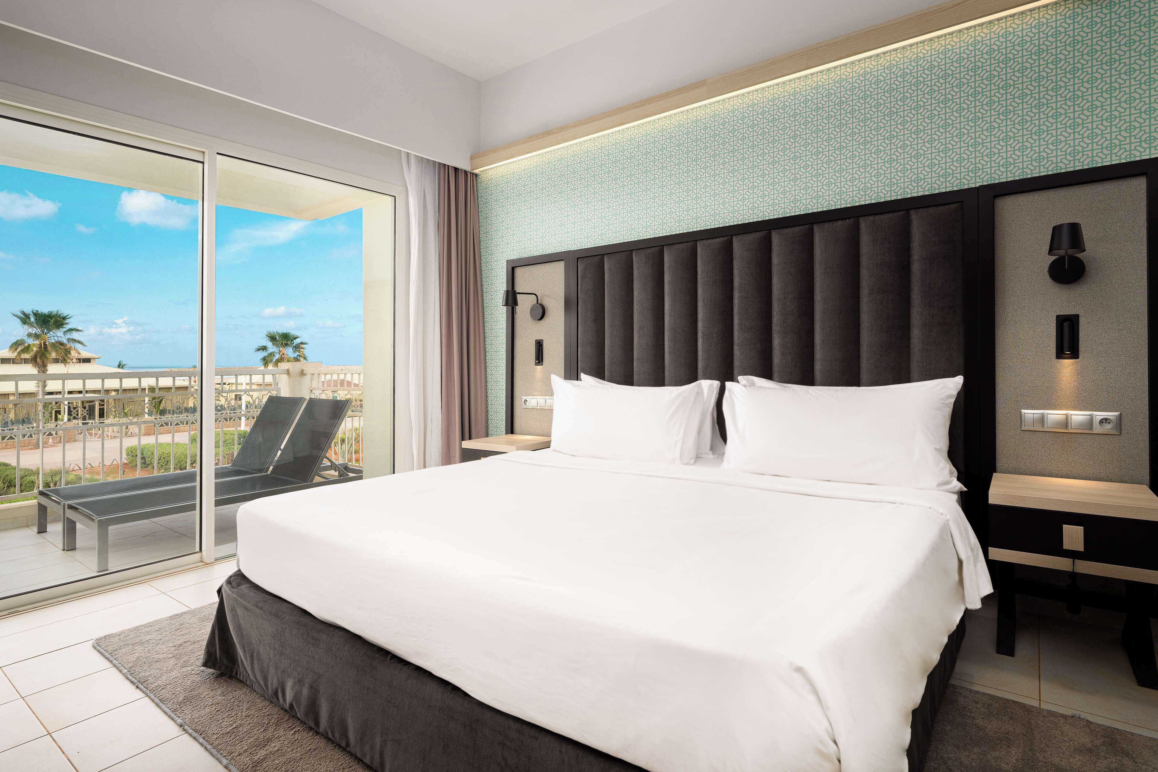 Habitación de hotel con una cama grande, una mesita de noche, una lámpara fijada a la pared y vistas a palmeras a través de una puerta corrediza de vidrio.
