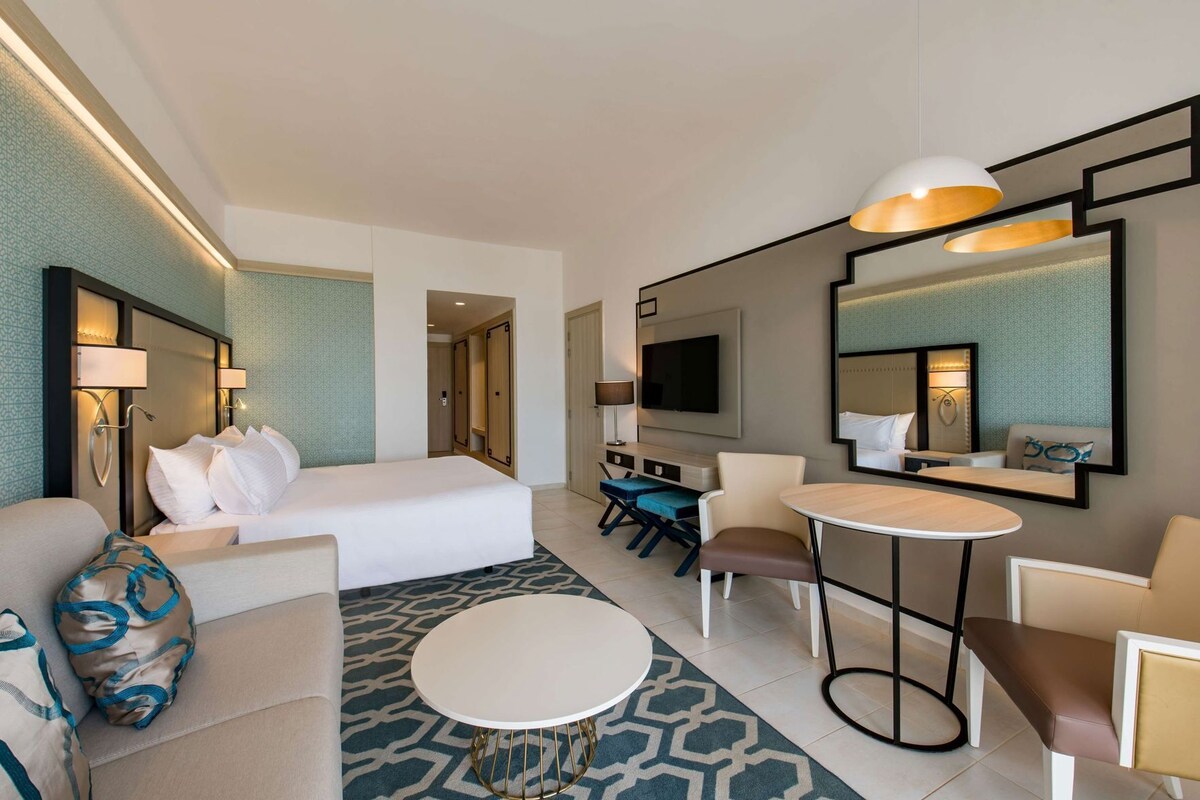 Una habitación de hotel con cama, sofá, mesa de comedor, televisor y espejo.