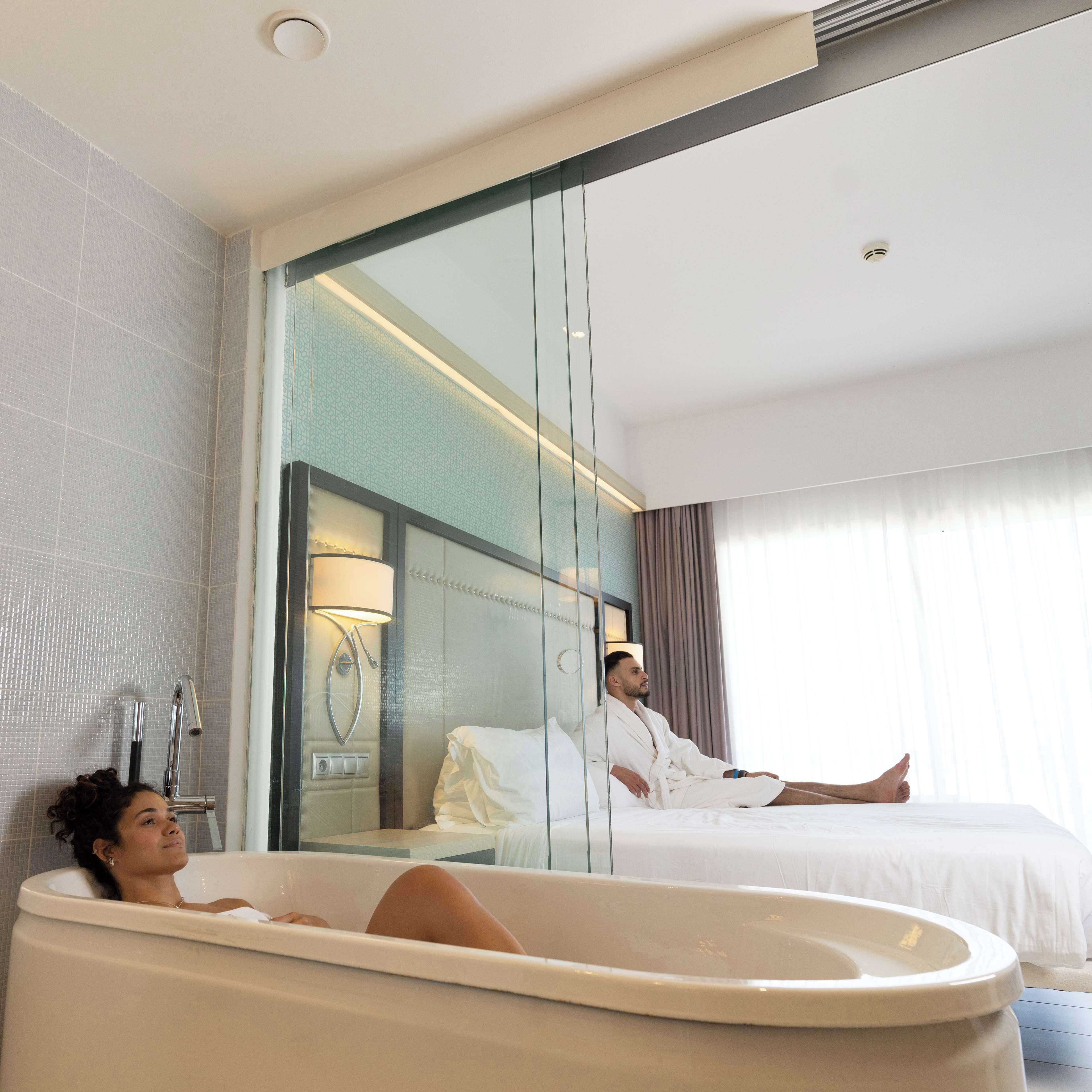 A modern hotel bathroom with a large bathtub, a glass shower enclosure, and a mirrored wall reflecting a person in a robe.