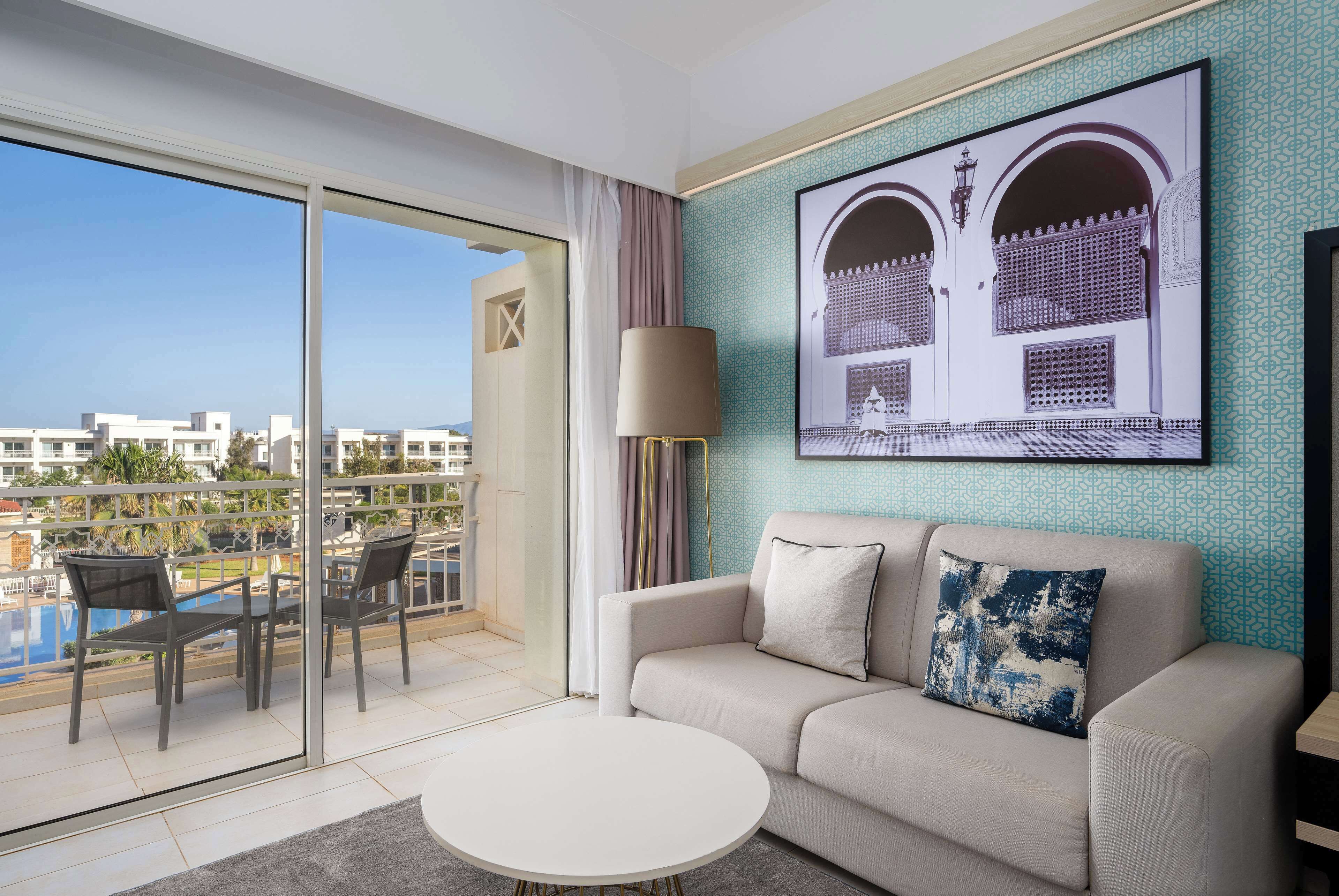 A modern living room with a sofa, a round coffee table, and a balcony with a pool view.