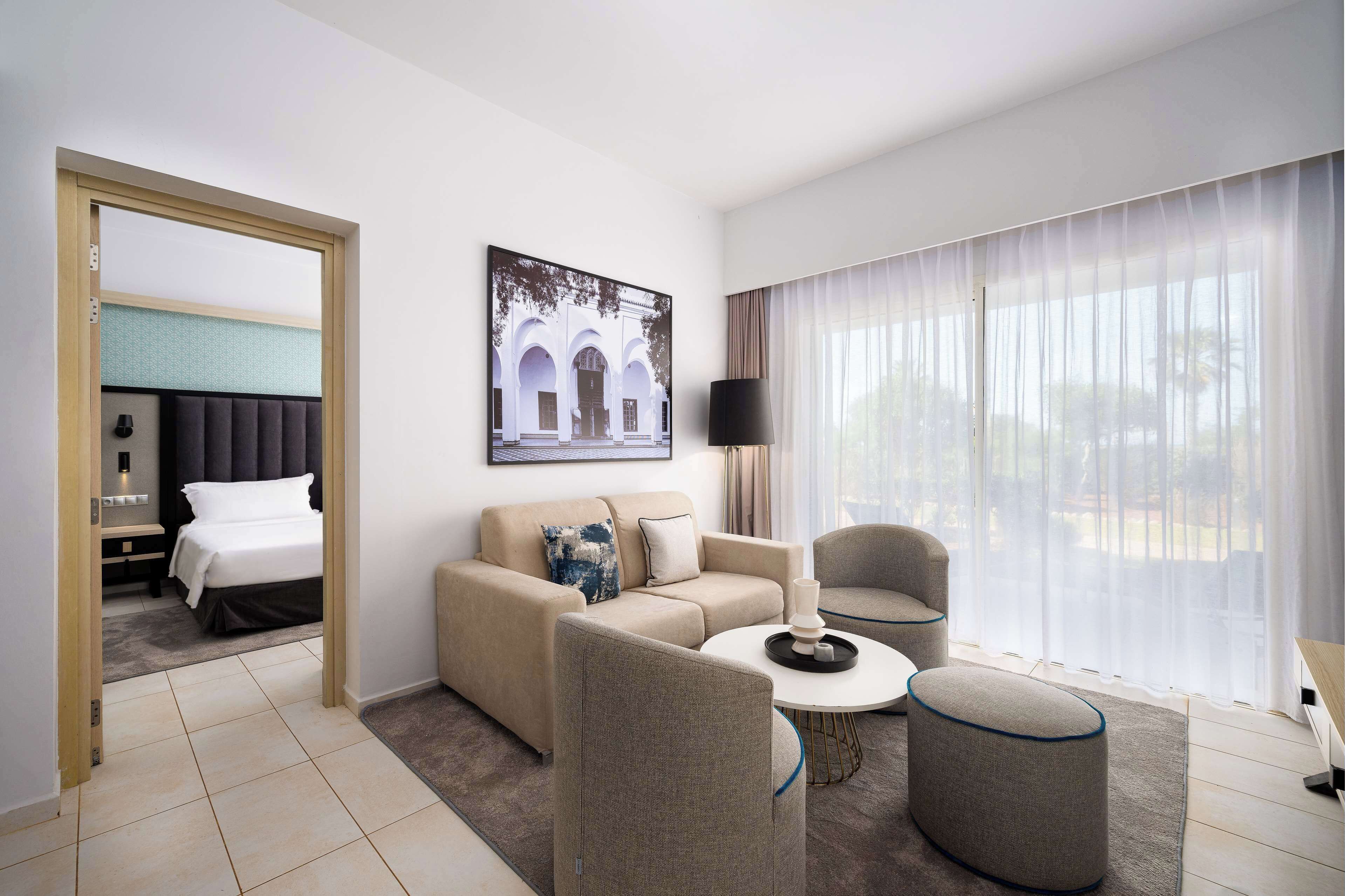 Una habitación de hotel con cama, sofá, mesita de centro y un gran ventanal con cortinas traslúcidas.