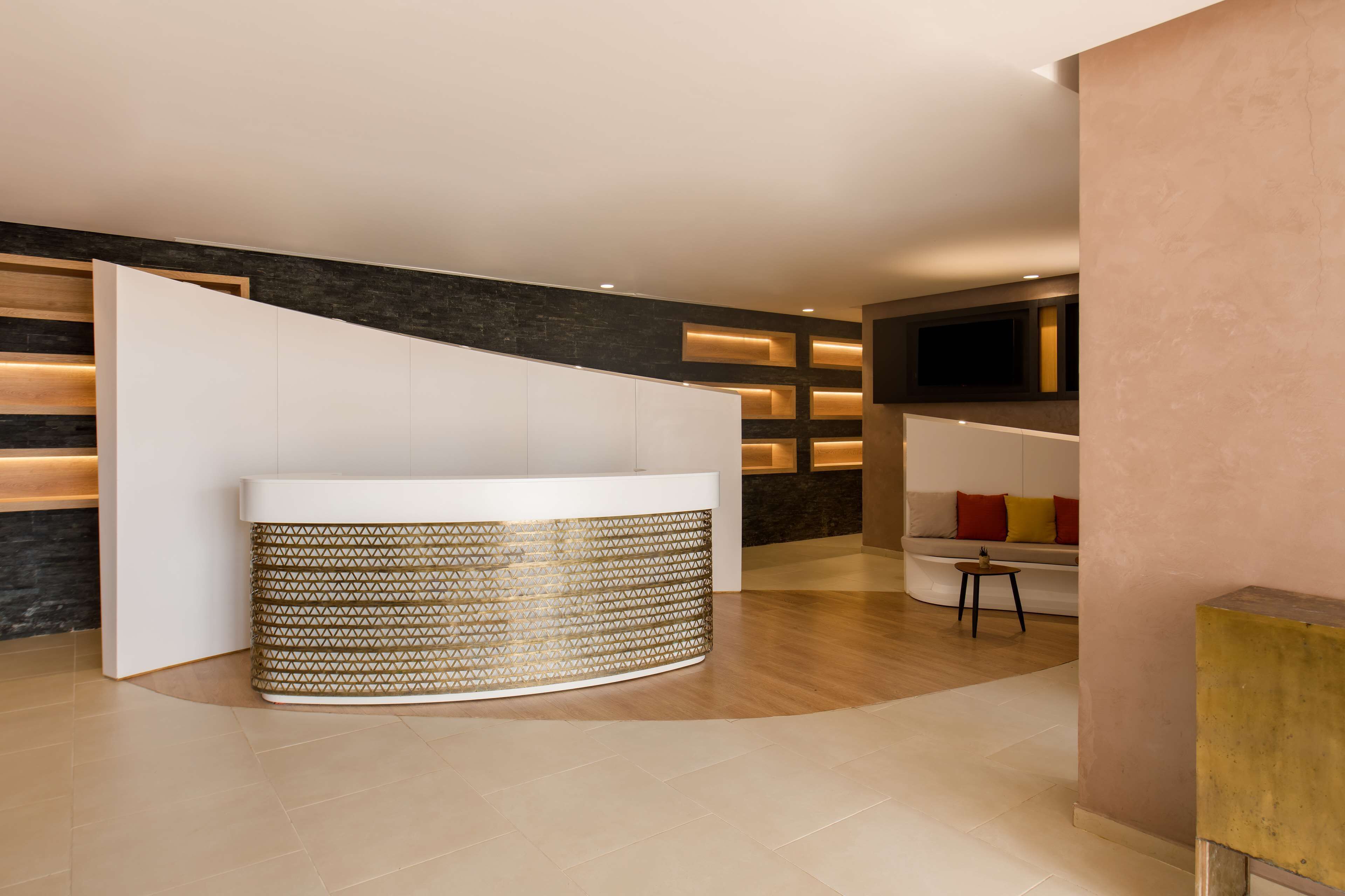 A reception area with a curved white counter, a patterned cylindrical base, a dark textured wall, and a seating area with cushions.