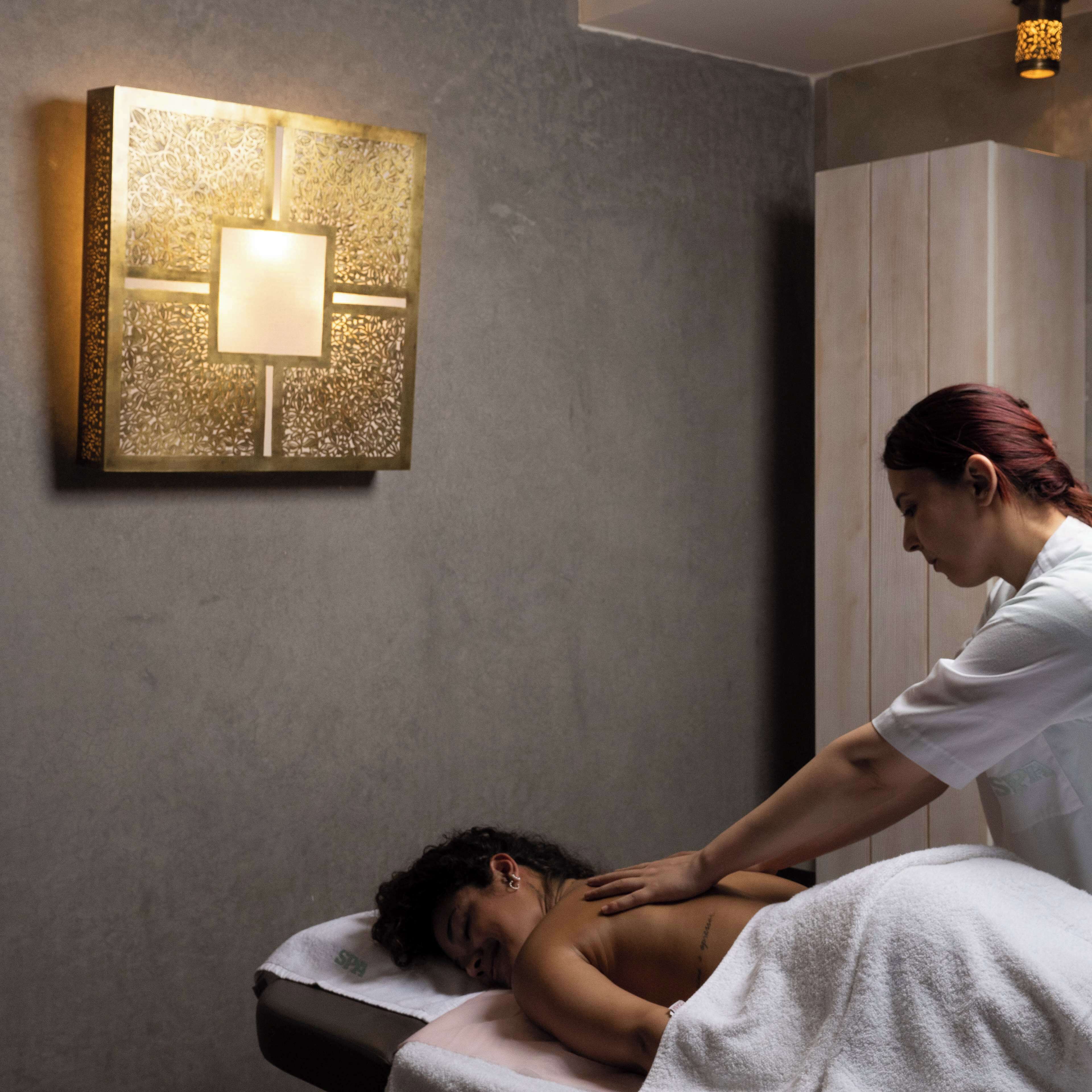 A massage therapy room with a massage table, a wall-mounted massage table lamp, and a spa towel on the floor.