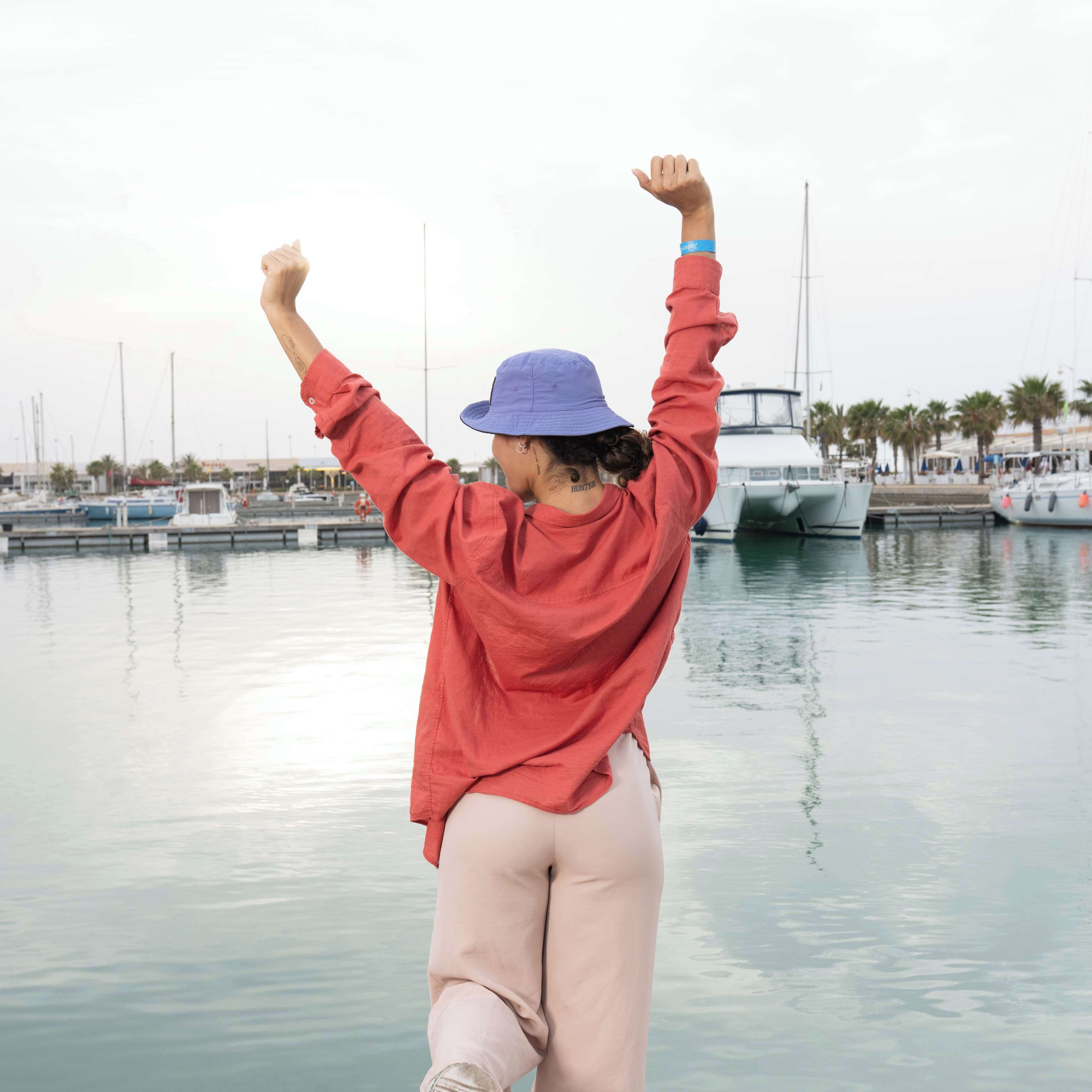 A person standing by a calm waterfront with boats and a marina in the background.