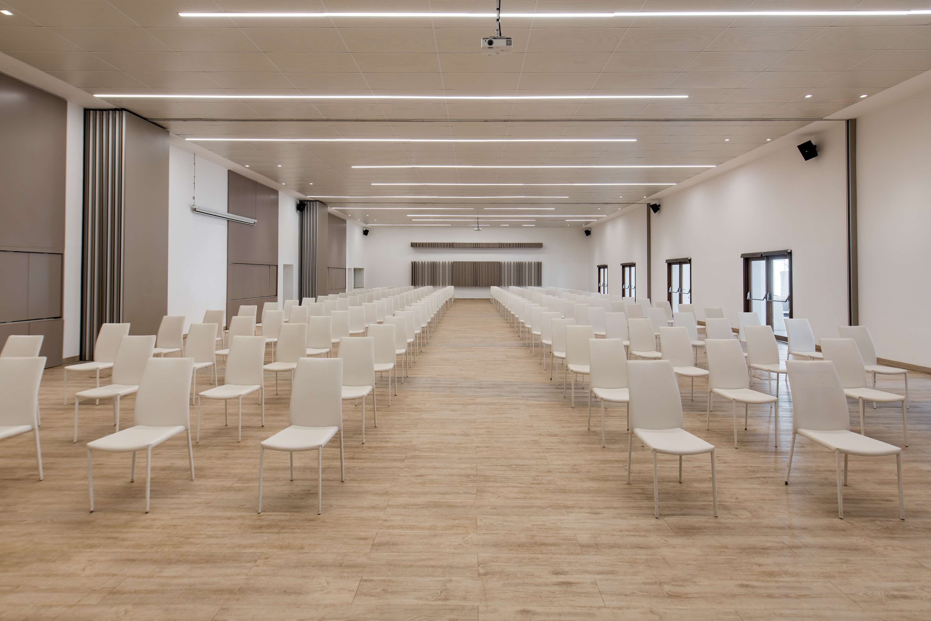Une salle de conférence avec des rangées de chaises blanches, un projecteur et une longue table à l’avant.
