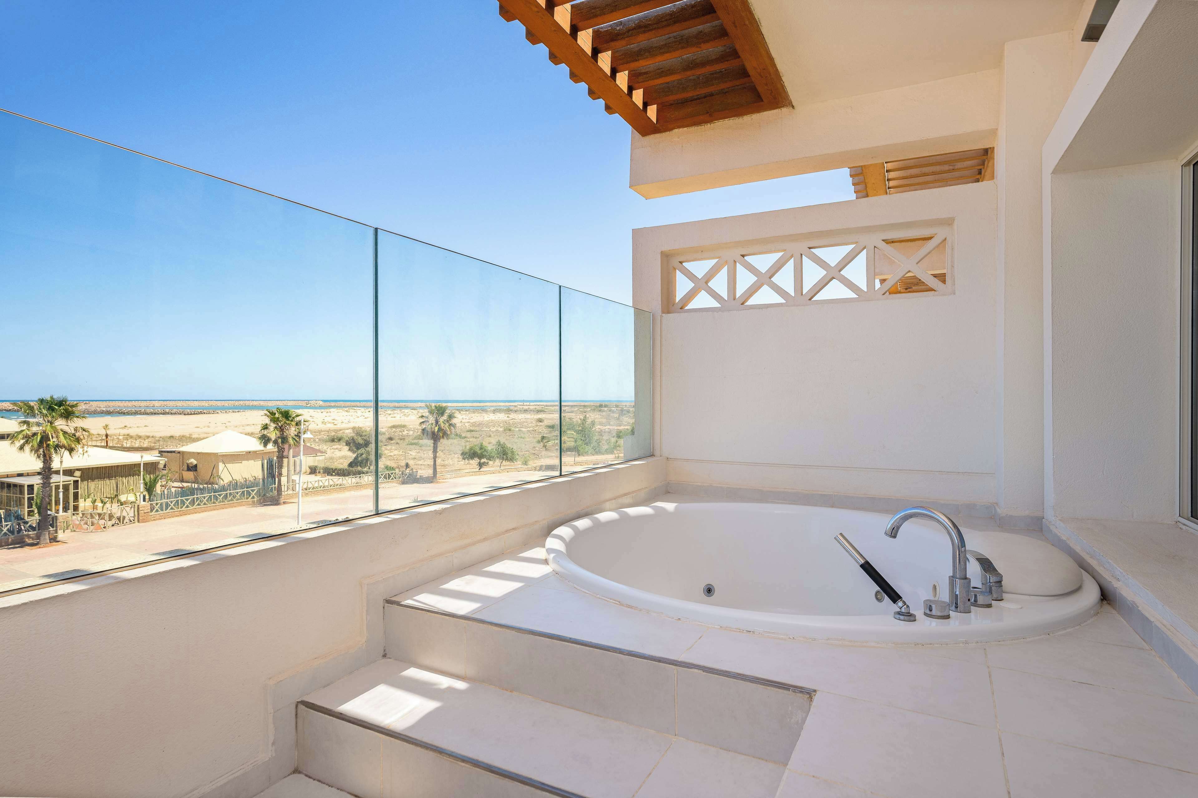 A modern bathroom with a large bathtub, a view of the beach, and a balcony with a glass railing.