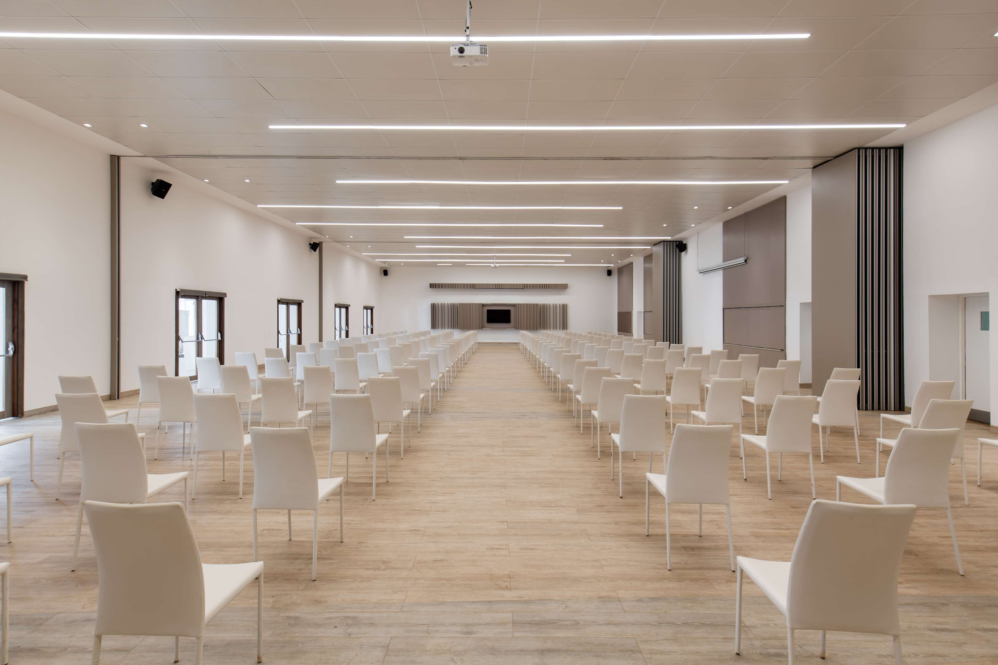 A spacious conference room with rows of white chairs, a wooden floor, and a modern ceiling design.