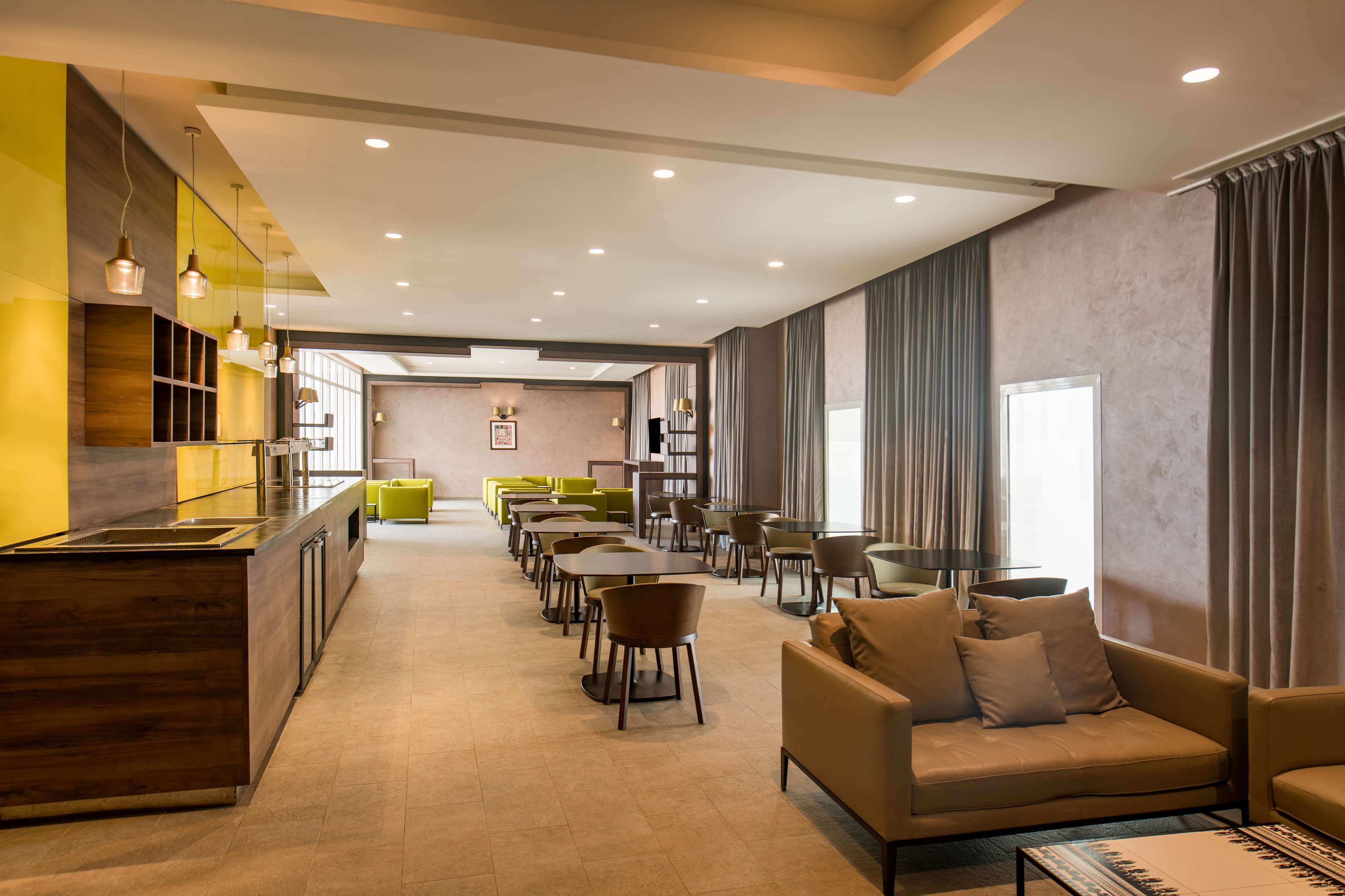A modern hotel lobby with a long reception desk, multiple seating areas, and a large window with curtains.