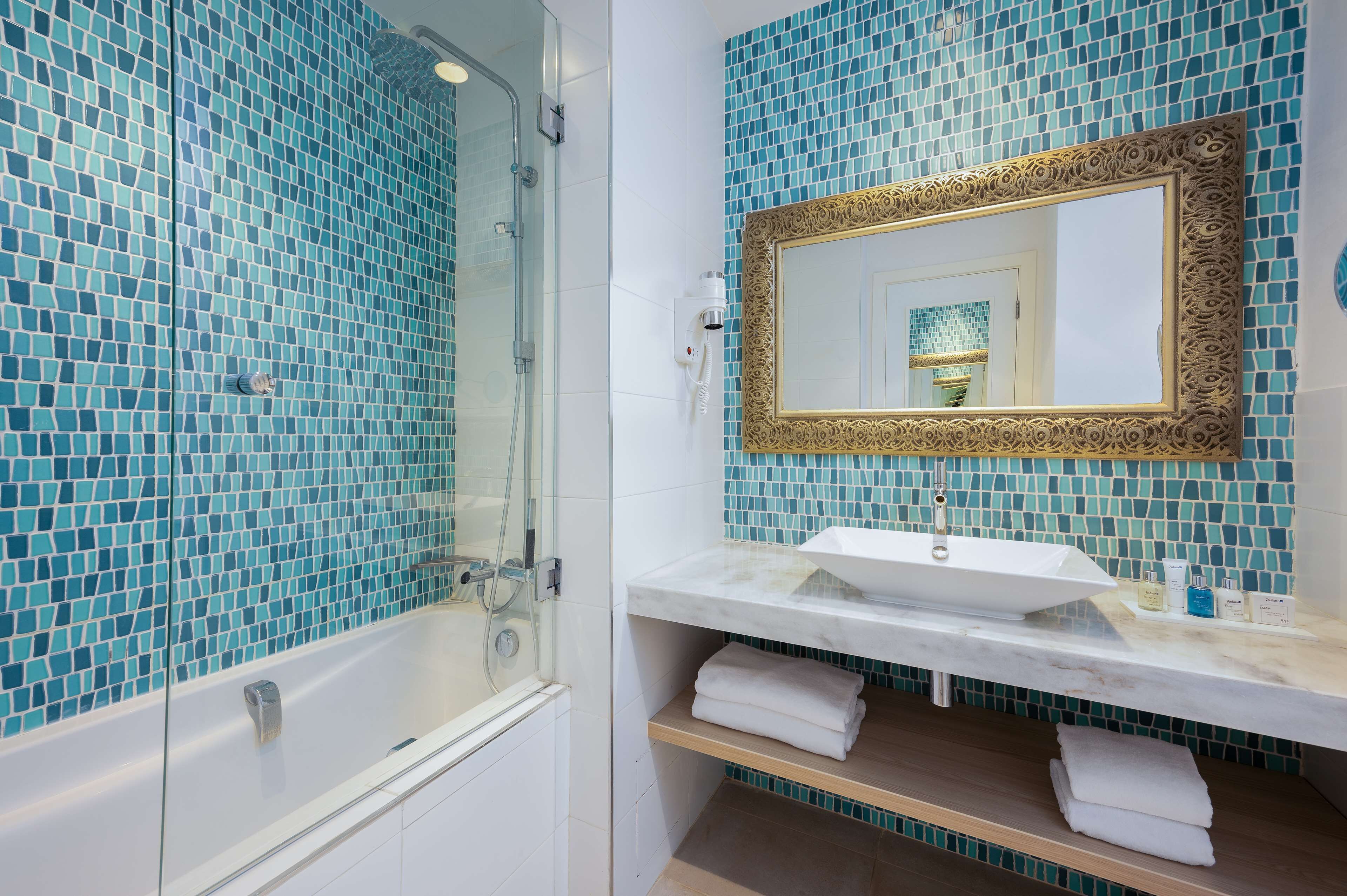 Une salle de bain dotée d’une douche à parois de verre, d’un lavabo blanc avec un plan de toilette en marbre et d’un miroir décoratif orné d’un cadre doré.
