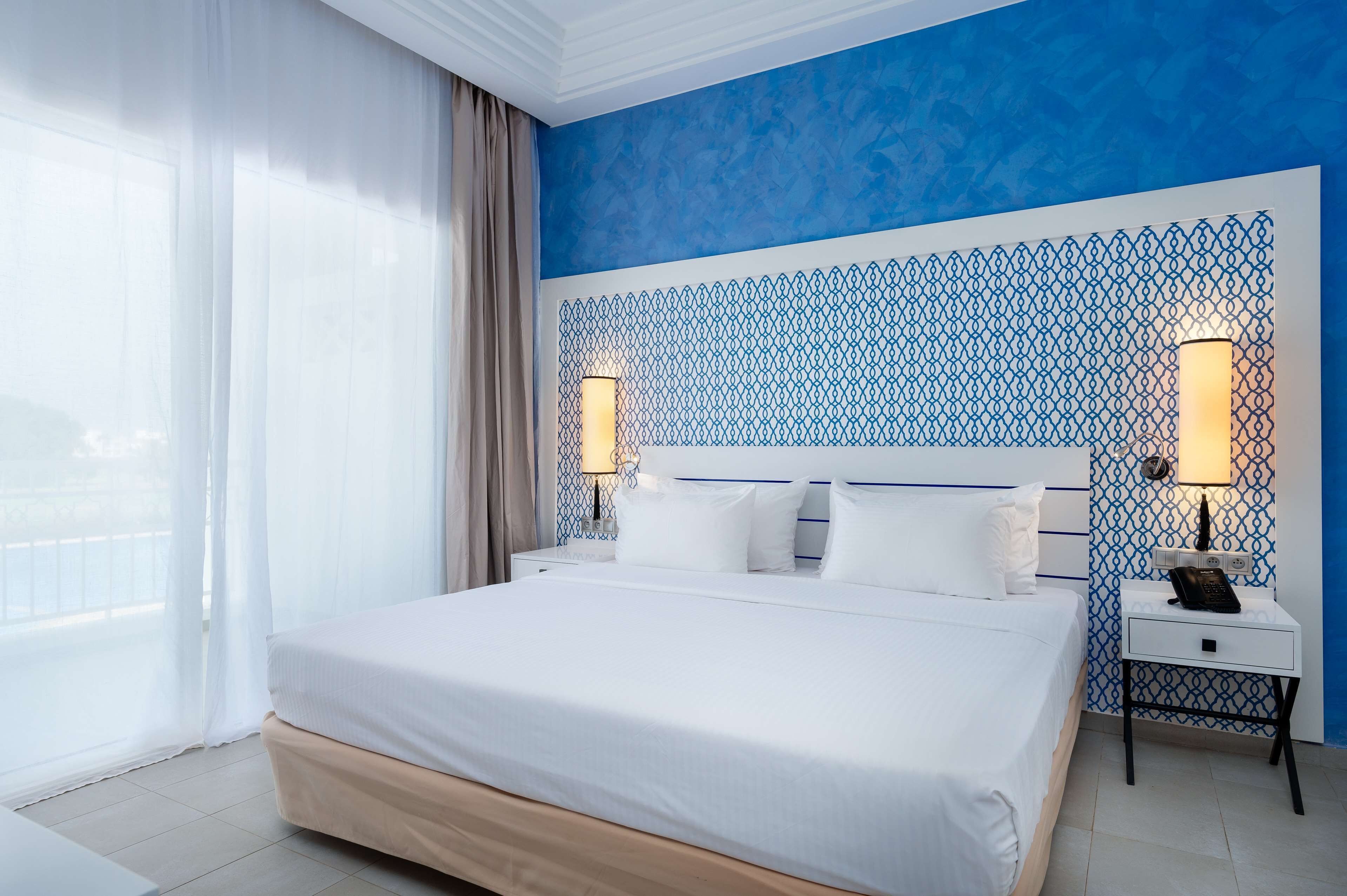 Habitación de hotel con una cama grande, dos mesitas de noche con lámparas, vistas a través de cortinas translúcidas y una pared con estampado azul y blanco.