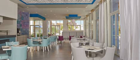 En rummelig restaurant med moderne indretning, hvide og turkise stole, et mønstret loft og store vinduer med udsigt til det fri.