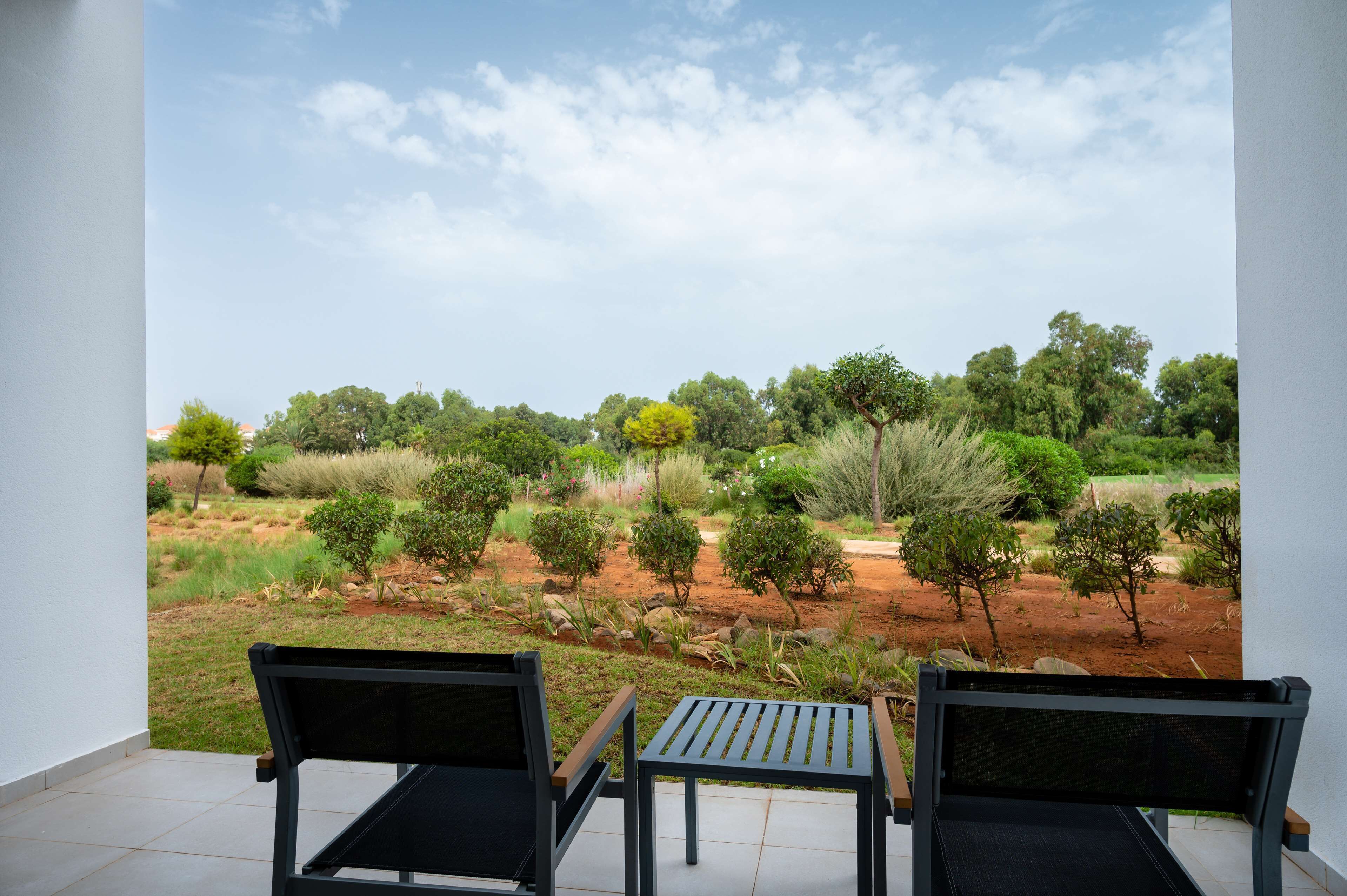 A balcony with two black chairs and a small table, overlooking a landscaped garden with various trees and shrubs.